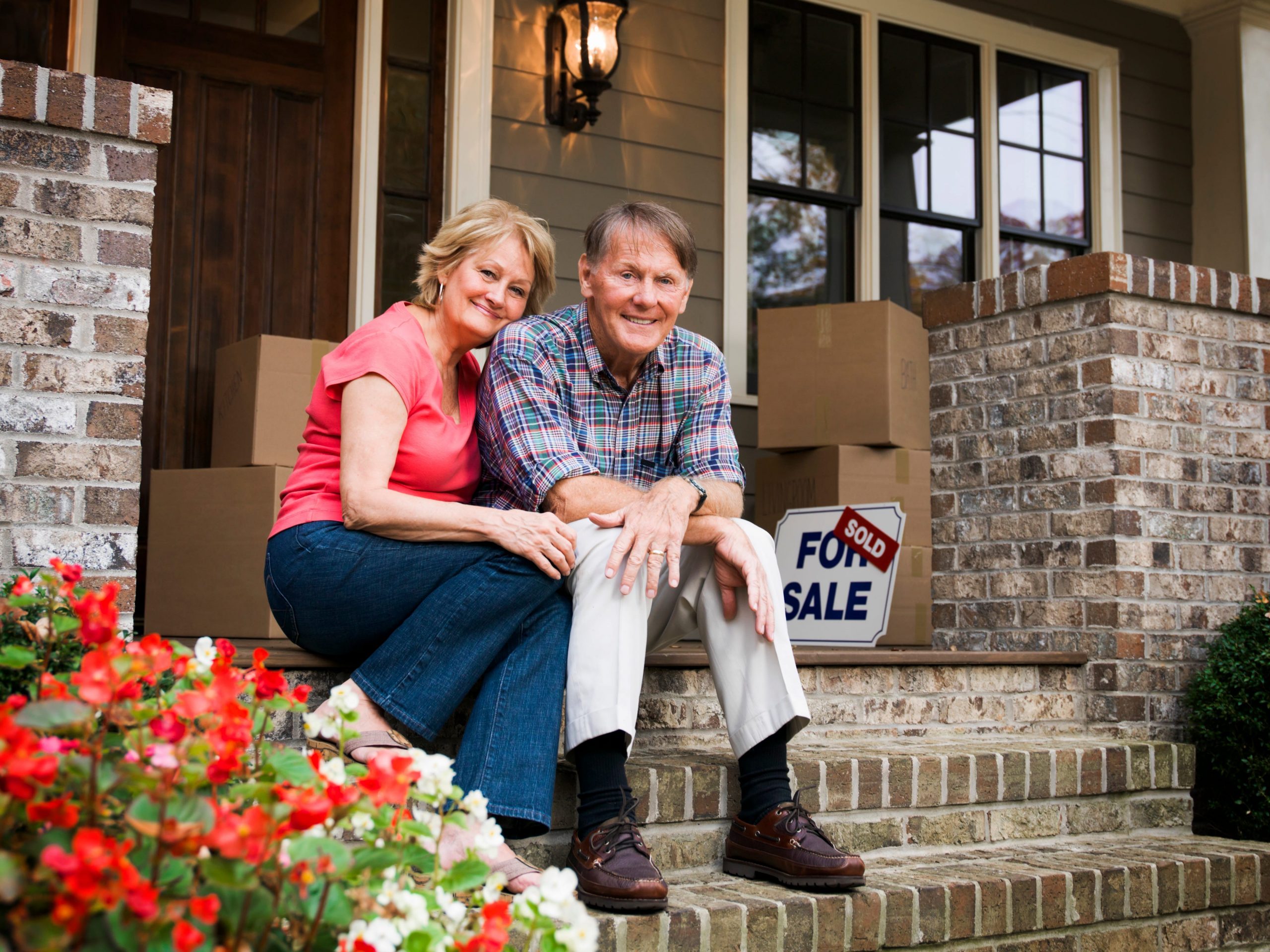 Couple sitting outside house with boxes and for sale sign