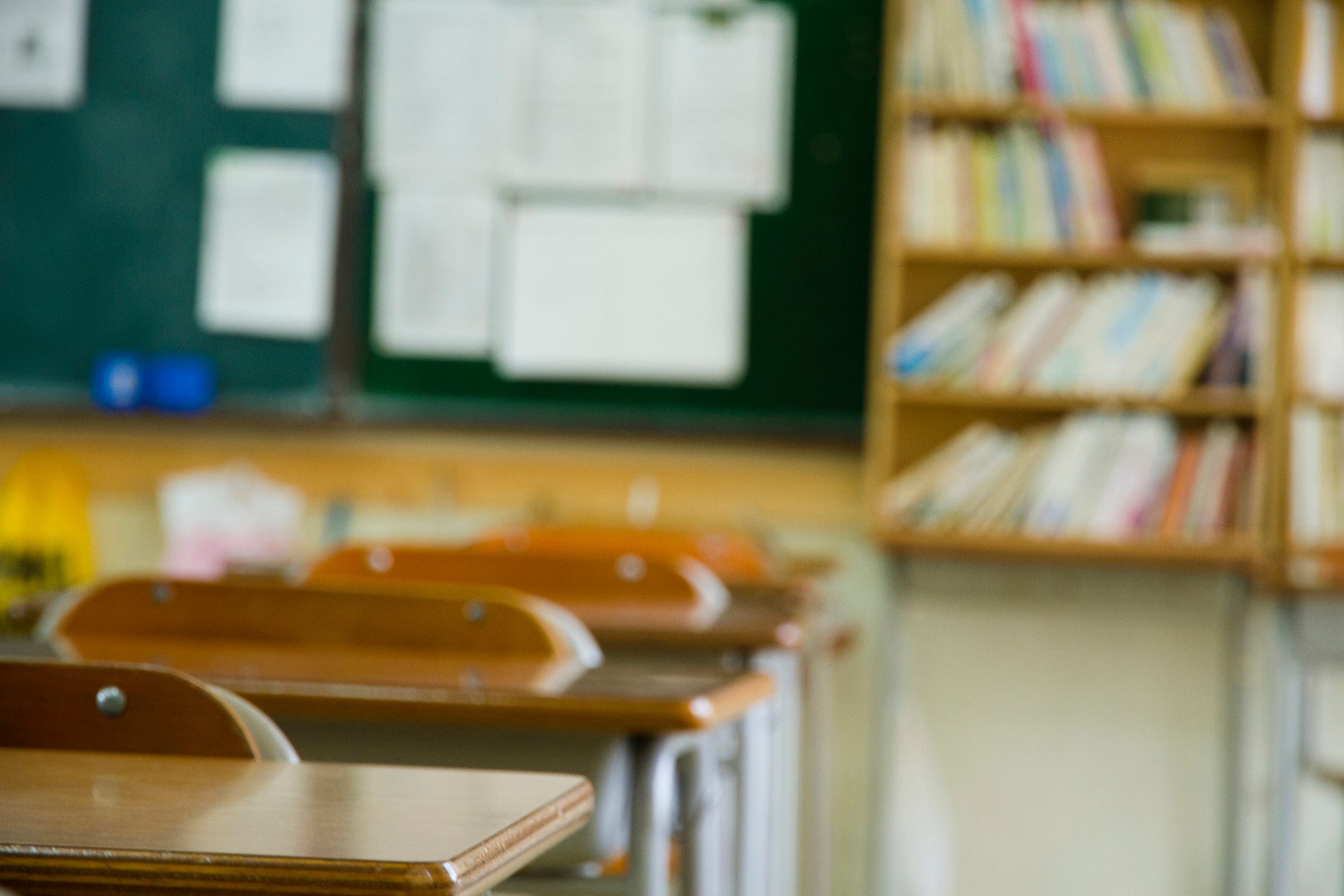 A school classroom, with desks, a blackboard, and bookshelves.