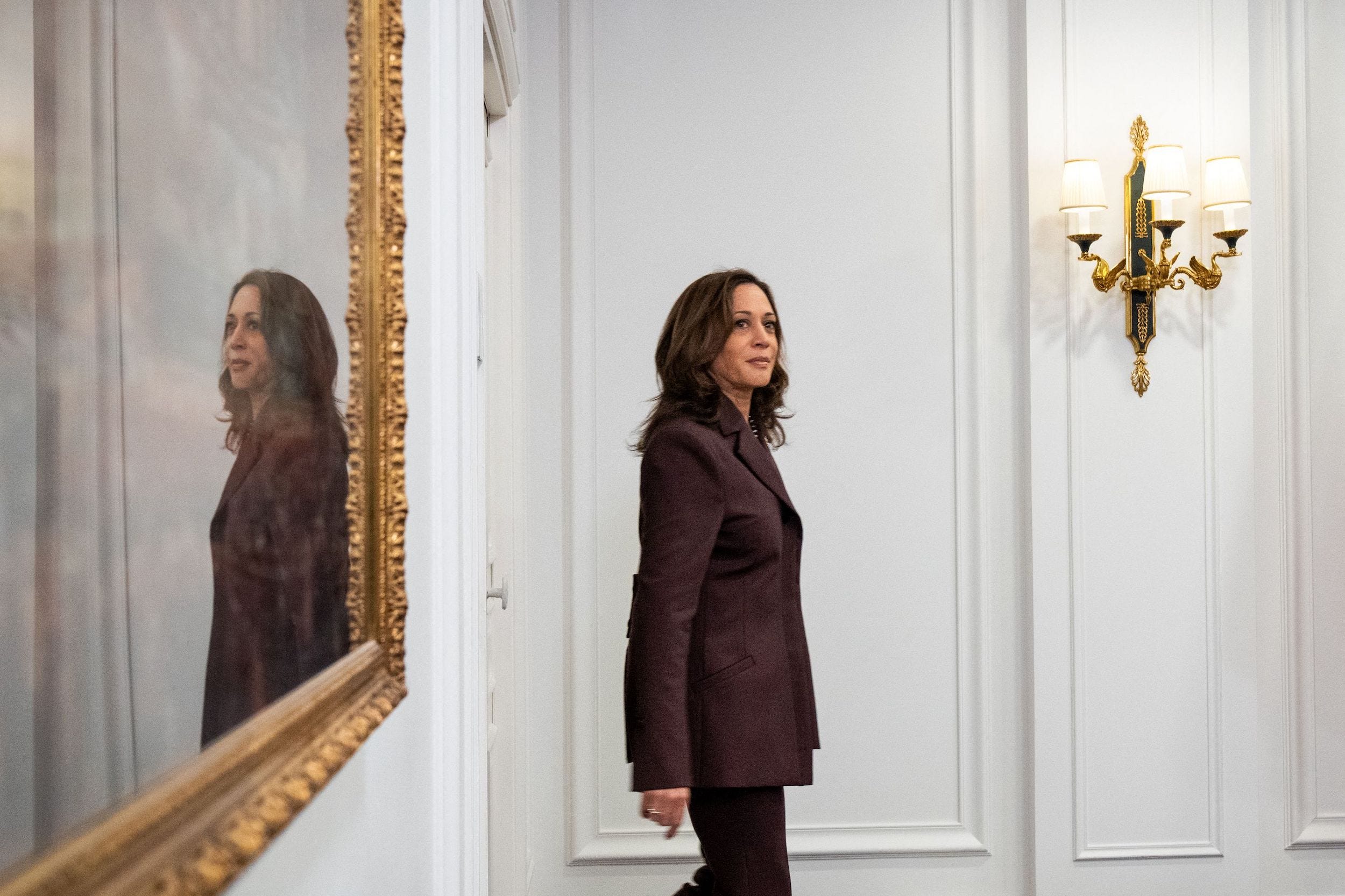 Vice President Kamala Harris walks into a room with her reflection on a mirror behind her.