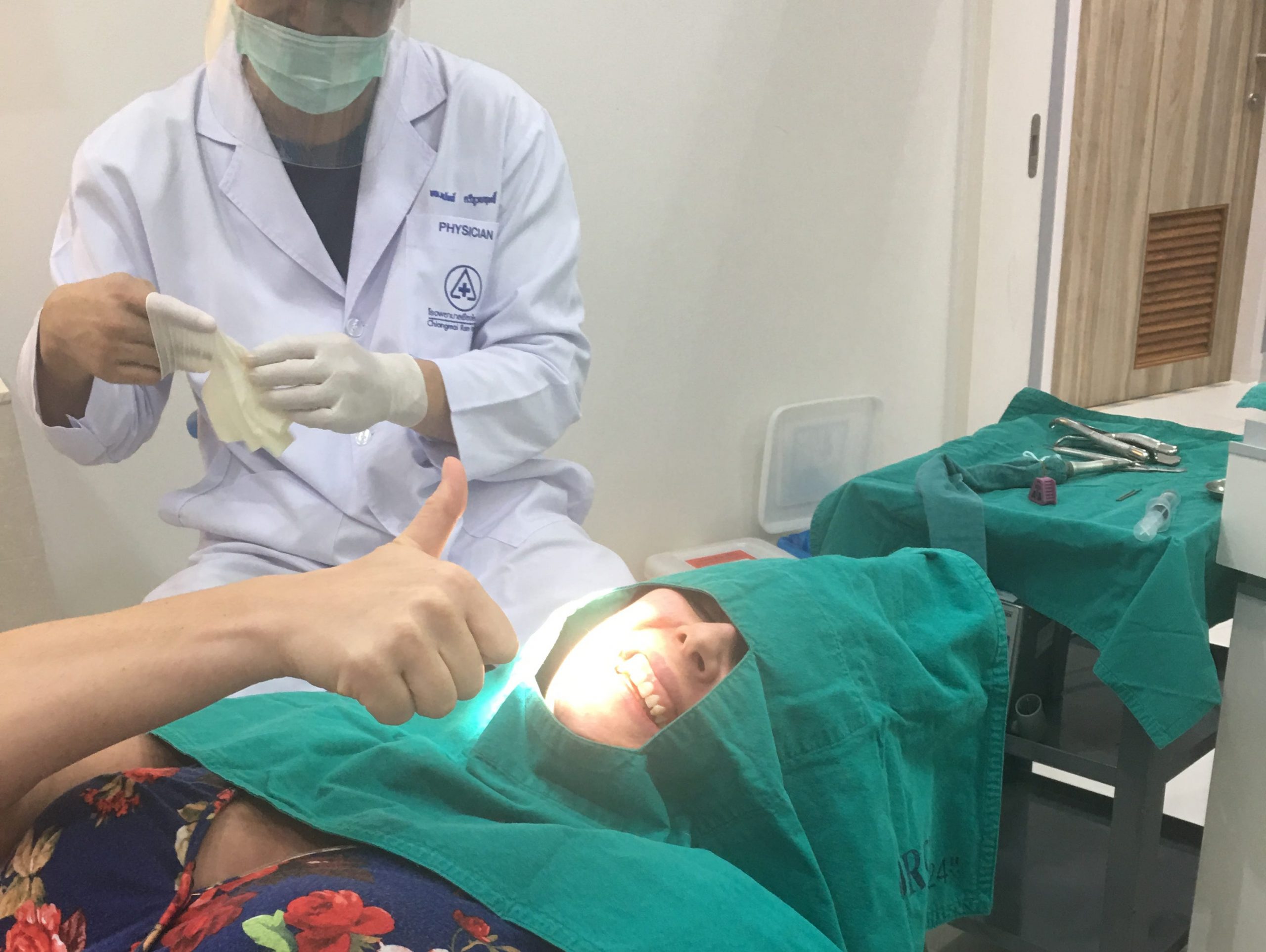 The writer giving a thumbs up under medical cloth while a dentist sits next ot her