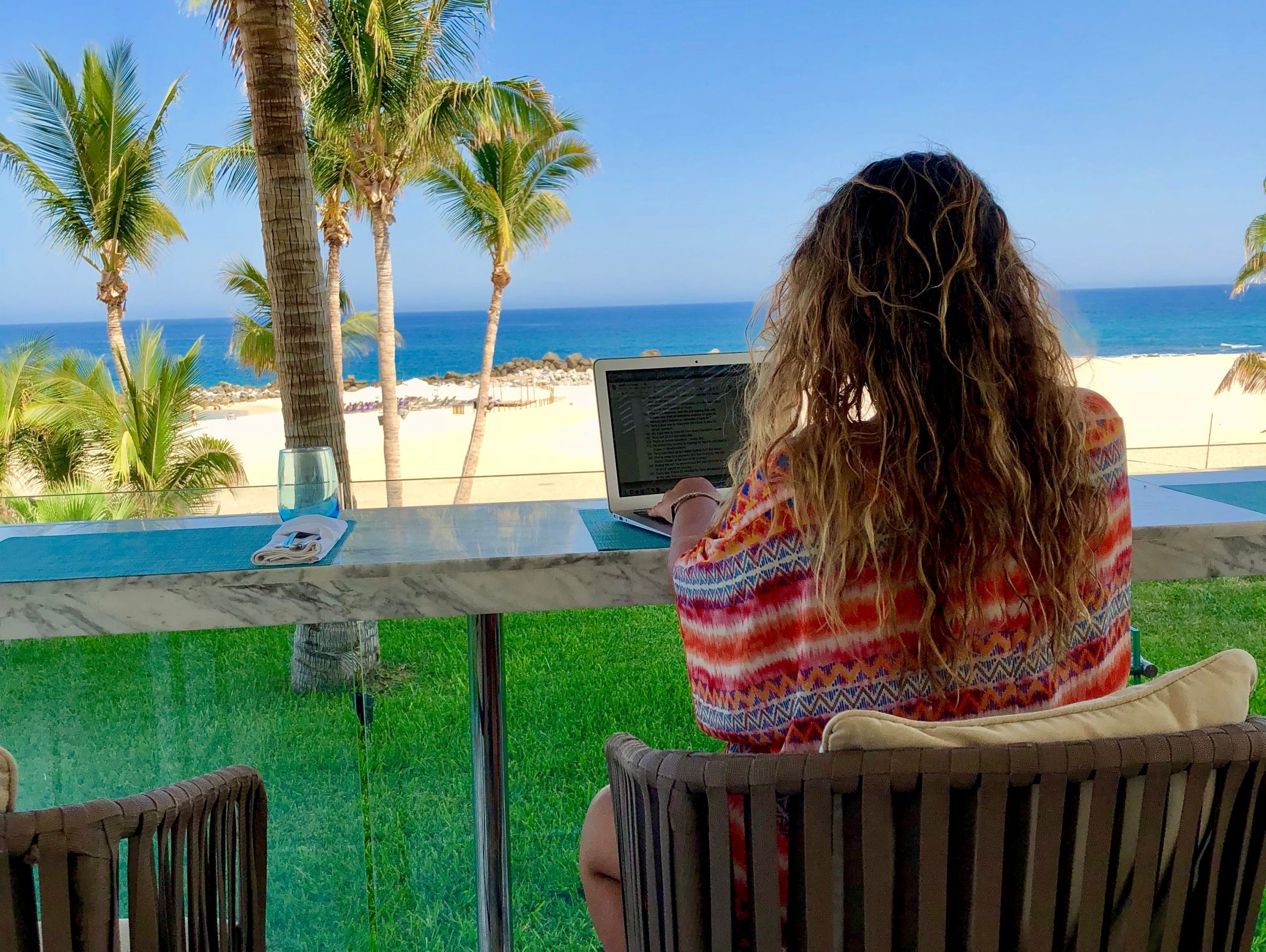 The writer sitting on a chair typing on her laptop and looking at the beach