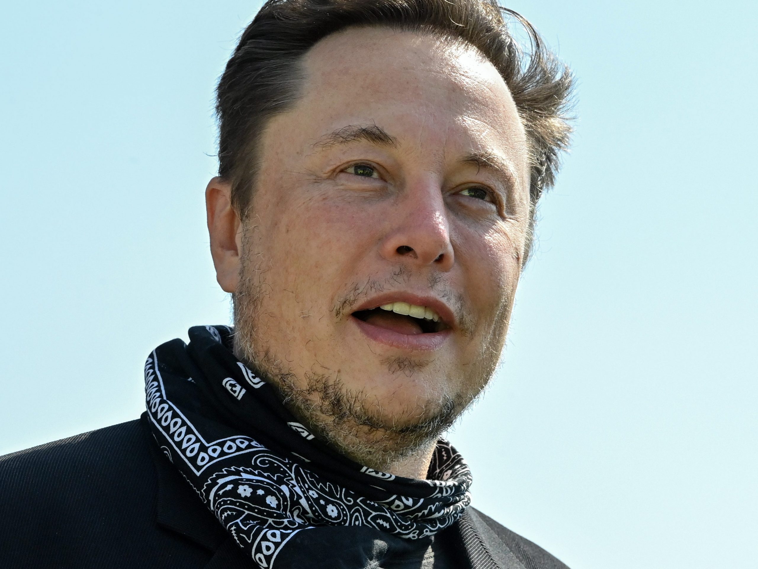 Tesla CEO Elon Musk wears a black and white bandana around his neck in front a light blue sky.