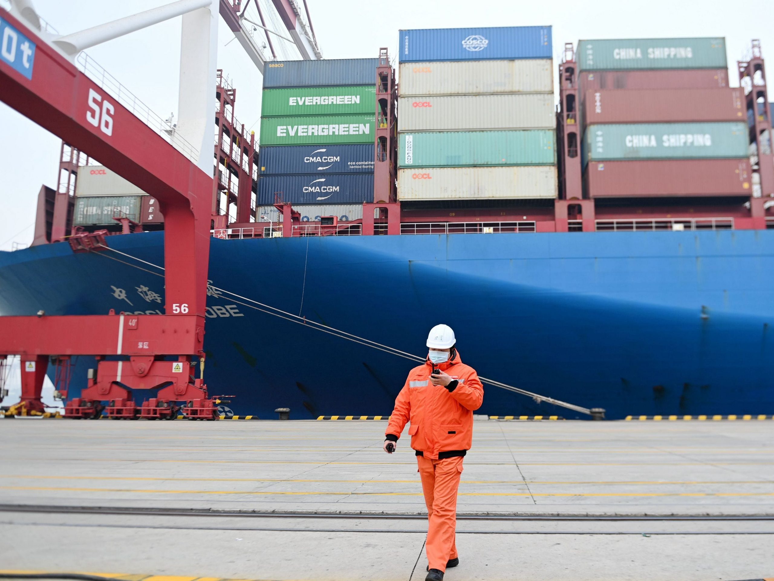 A masked man in orange in front of the side of a container ship in China.