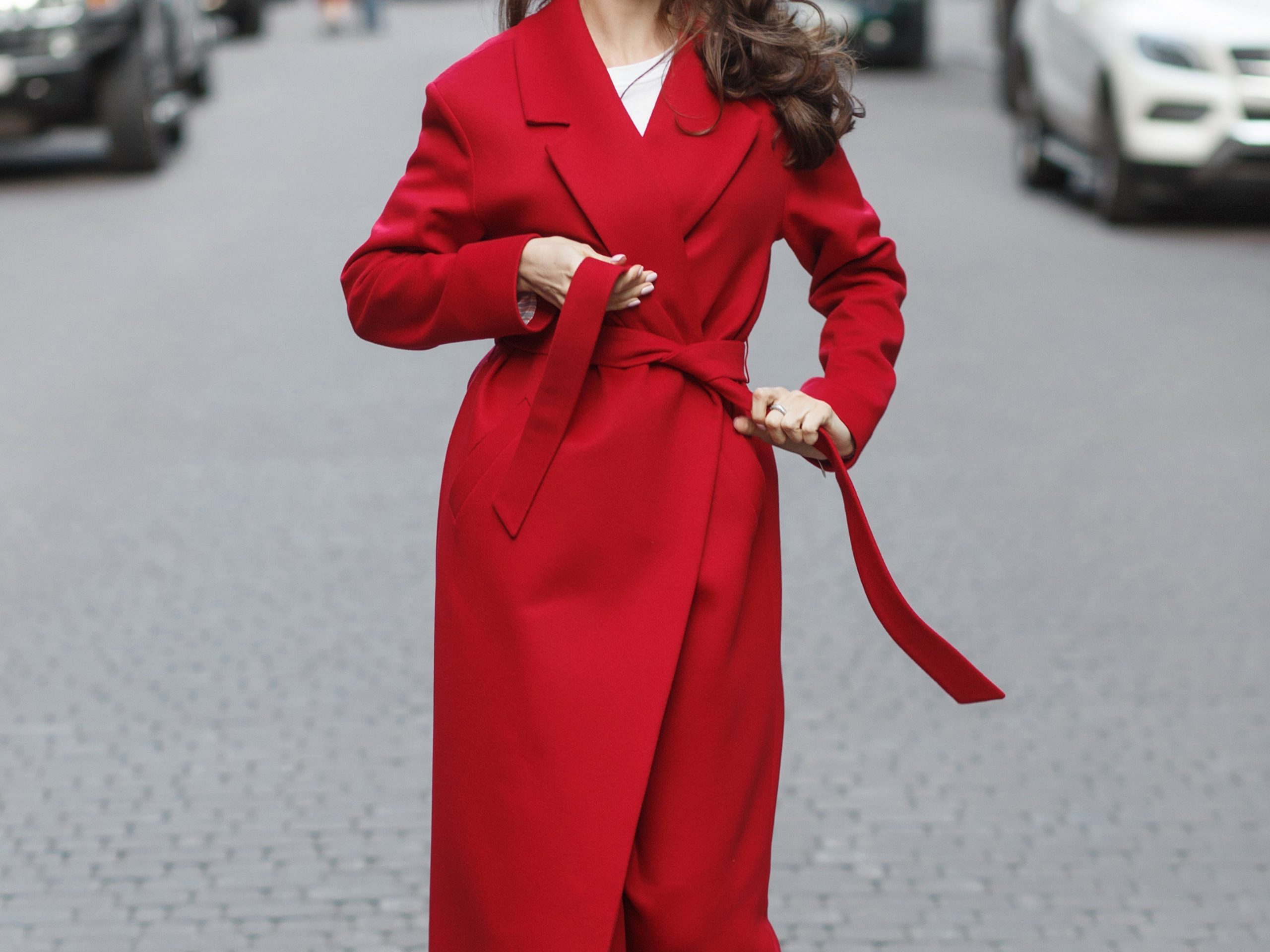 woman walking down a street tying the belt of her red trench coat
