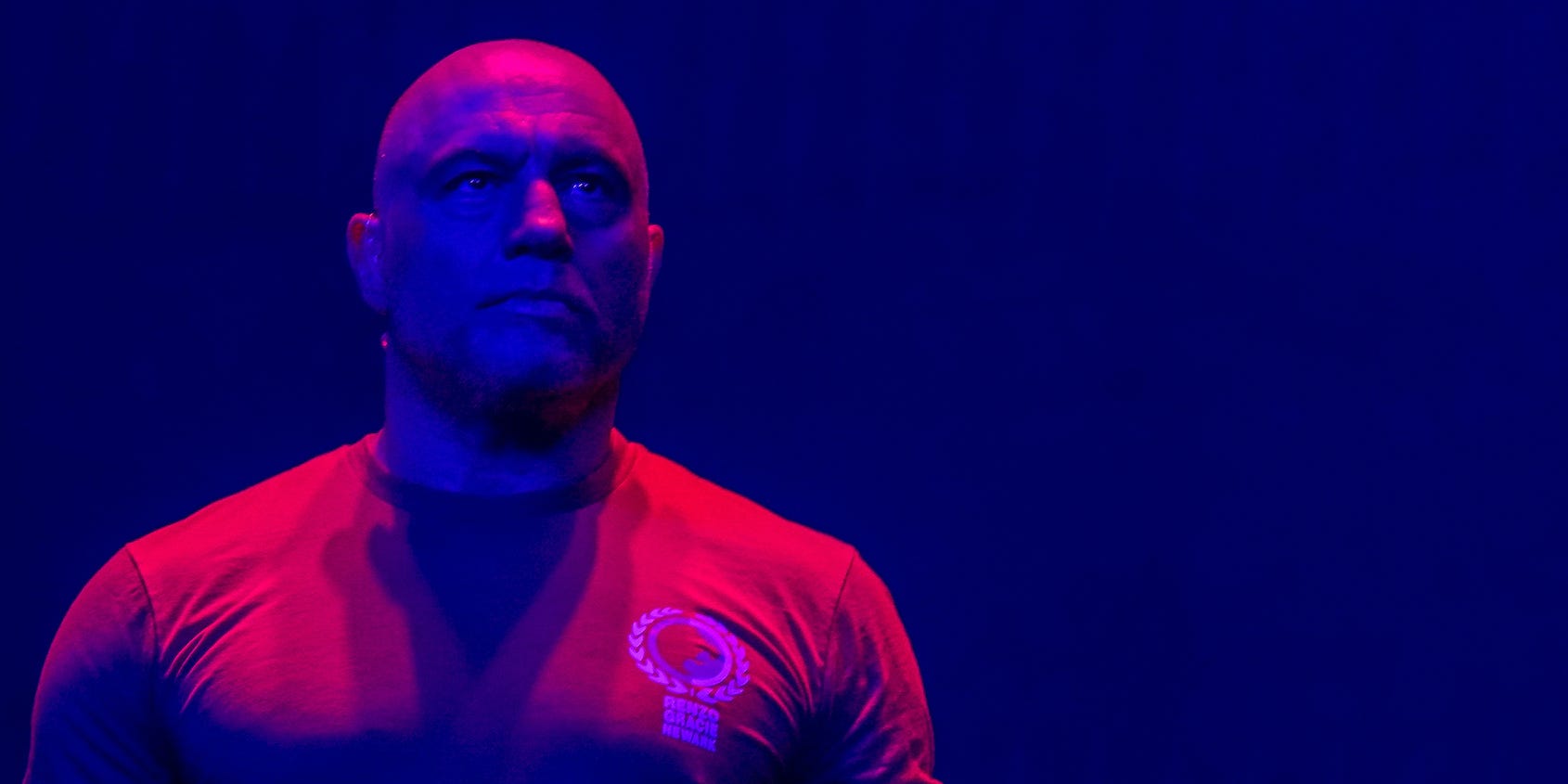 Drenched in a mix of red and purple light, Joe Rogan stands expressionless.