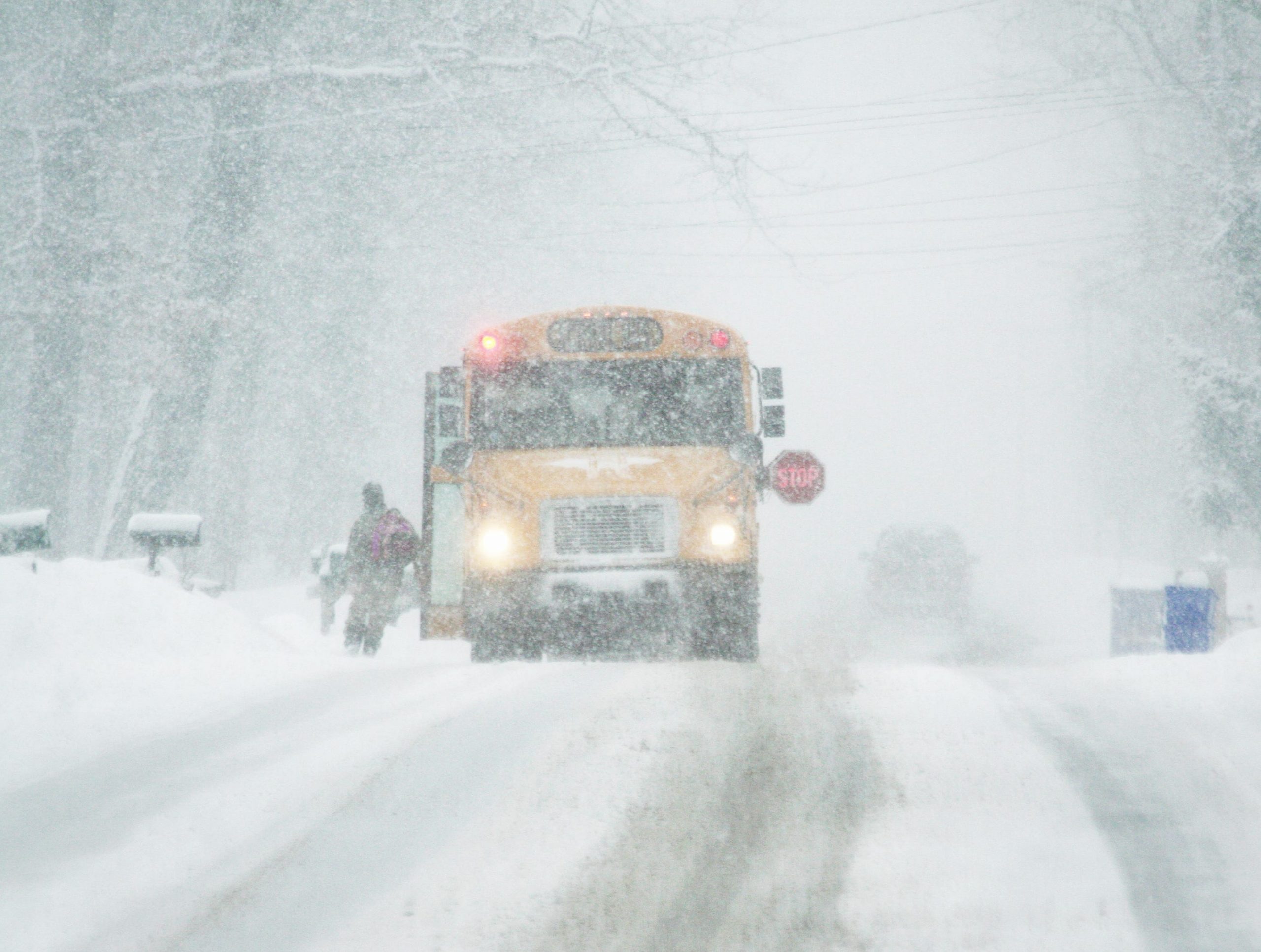 A yellow school bus with open door, flashing lights and outstretched warning stop sign in a raging snow blizzard is waiting for bundled up child who has just run across the road to board.