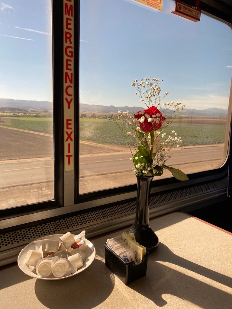 a rose in front of a dining car window next to a box of sugars and bowl of cream