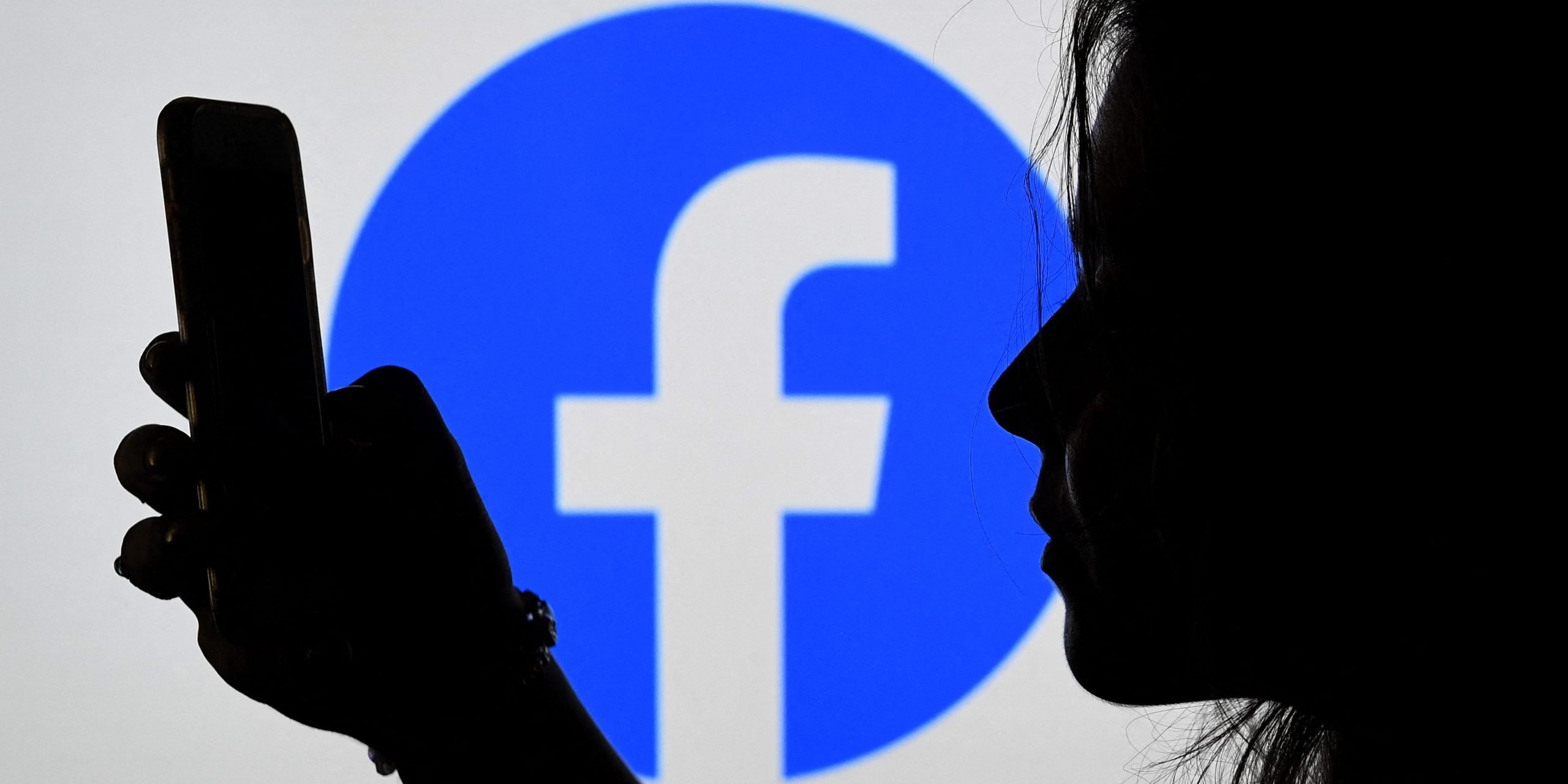 A person looks at. a smart phone with a Facebook logo displayed in the background