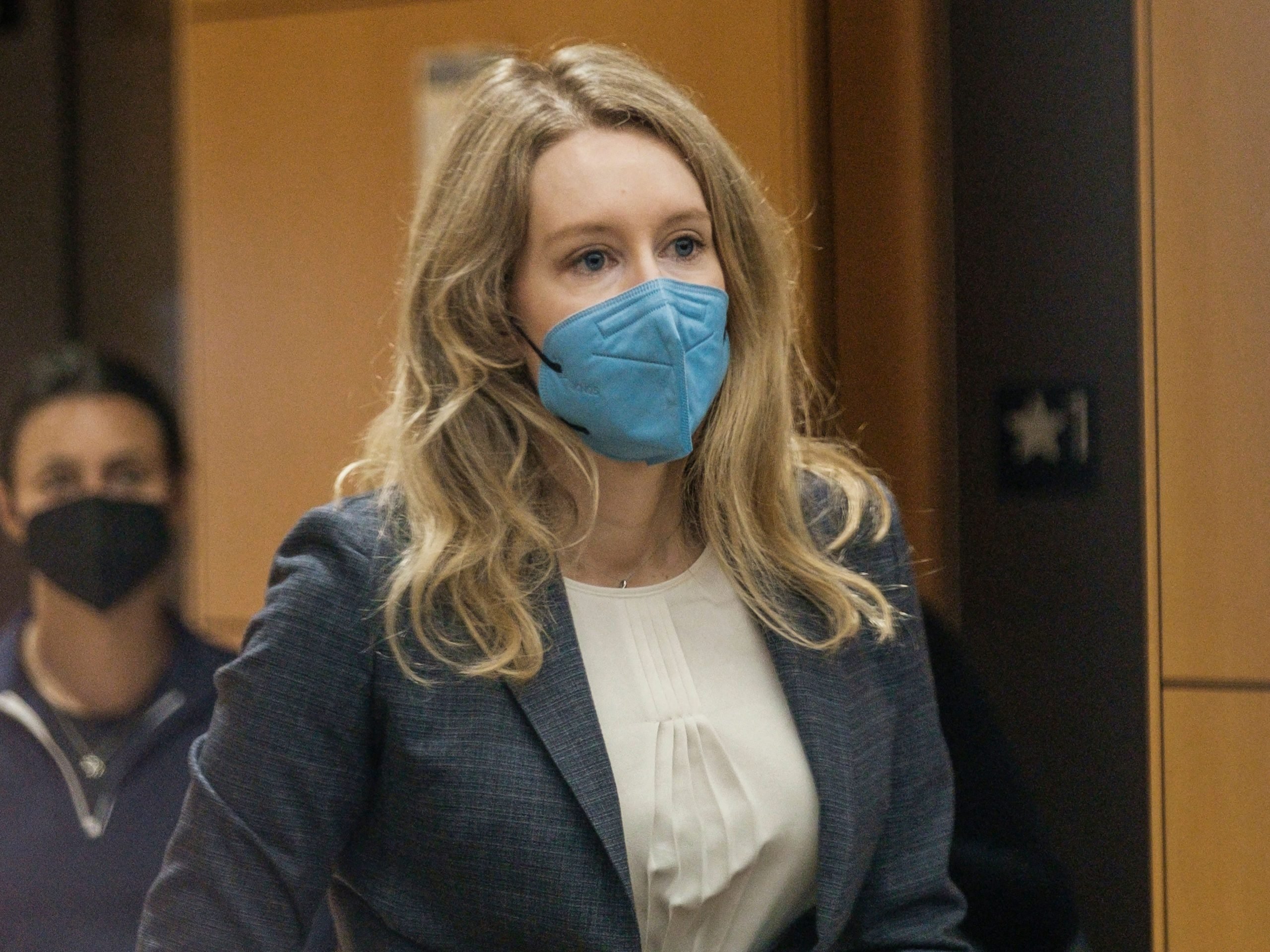 Theranos founder Elizabeth Holmes looks ahead while wearing a blue mask as a man stands behind her