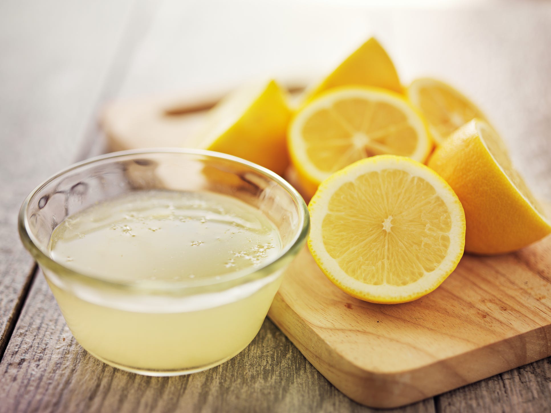 Lemon halves sitting on a wooden cutting board next to a glass bowl of lemon juice