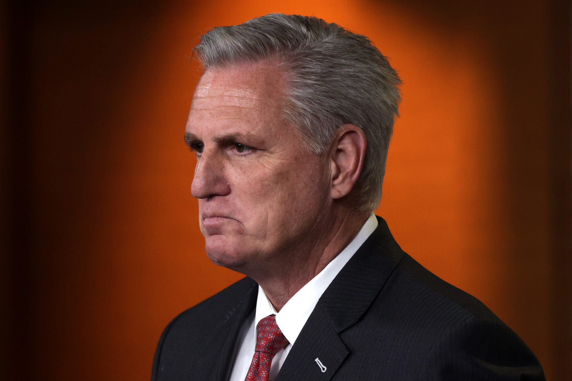 House Minority Kevin McCarthy, his lips turned down, stands against an orange background while wearing a dark suit, white shirt, and patterned tie.