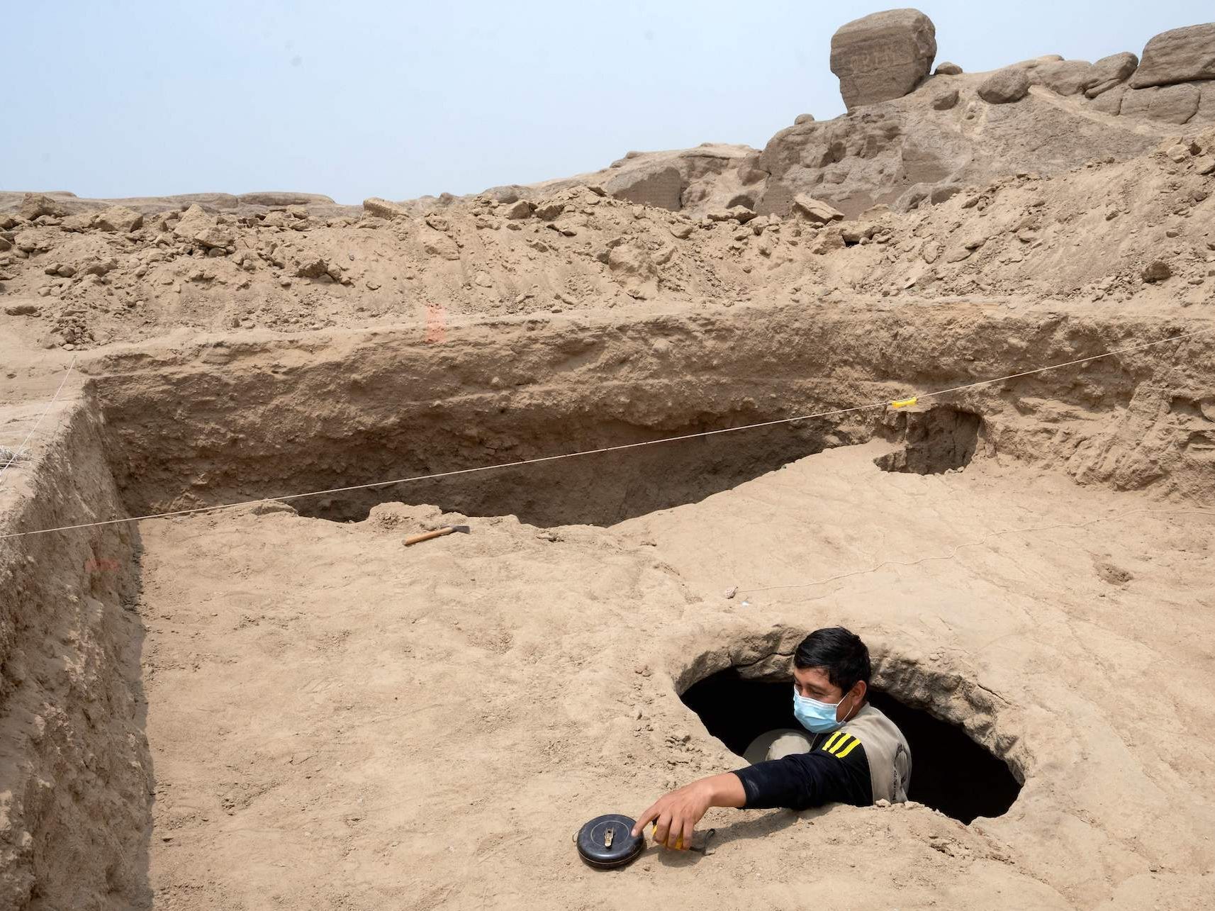 A man wearing a mask is seeing coming out of a hole drilled into the underground burial chamber in which the mummy of Cajamarquilla was found.