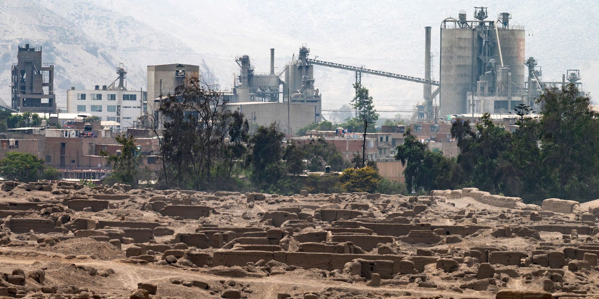 A view of Cajamarquilla, an archeological site outside of Lima, Peru, shows the excavated landscape in front of a backgorund of industrial buildings.