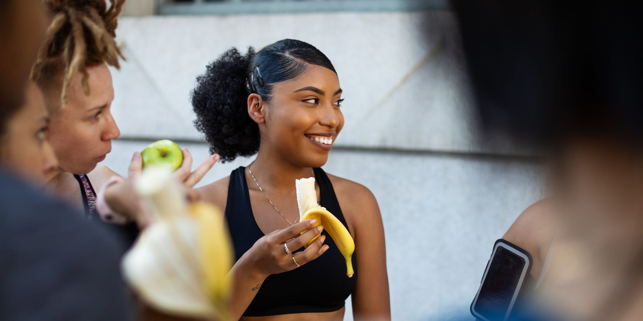 A Black woman in workout attire holds a half-eaten banana.