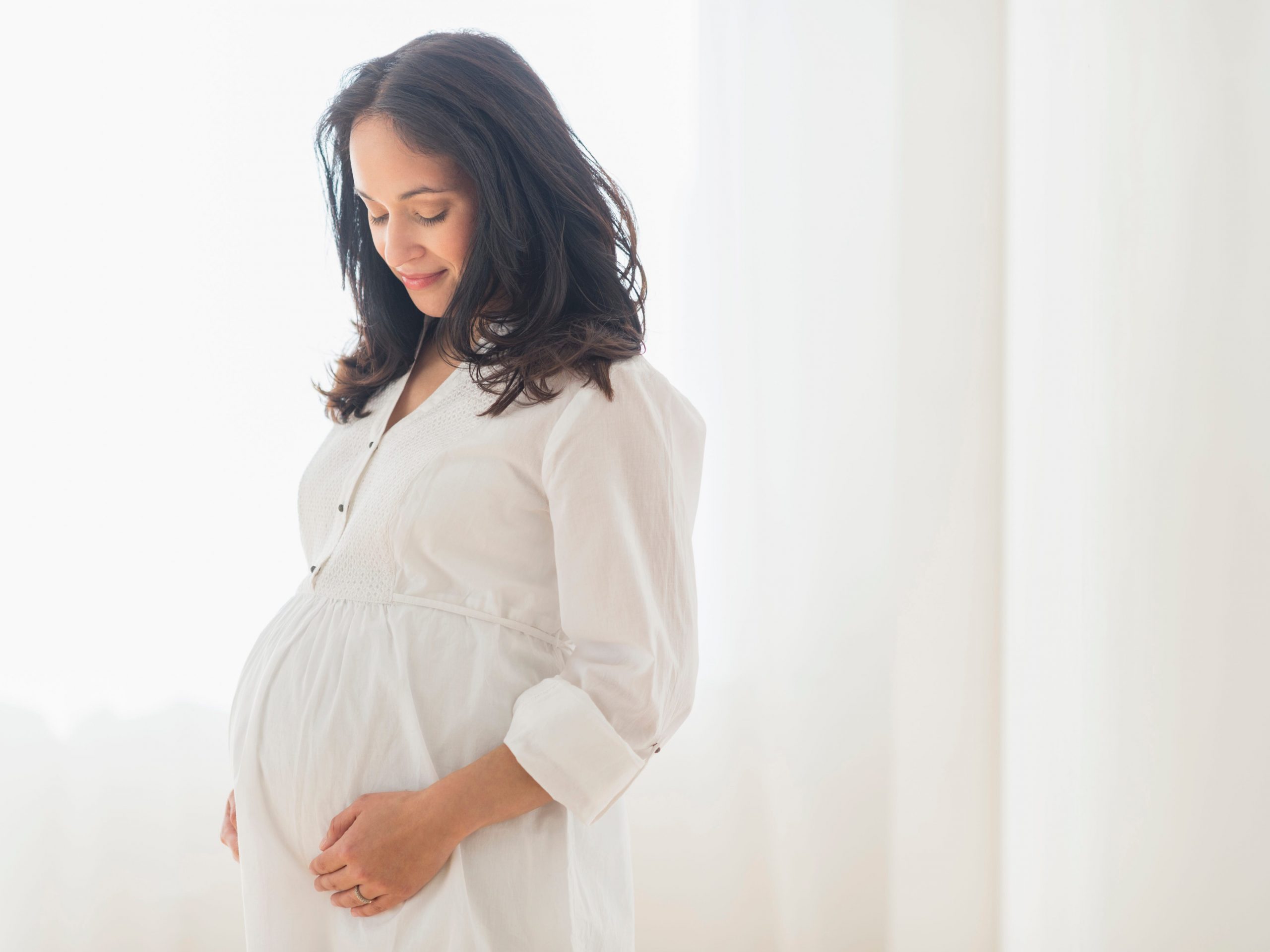 A pregnant woman wearing white and placing her hands on the sides of her stomach.