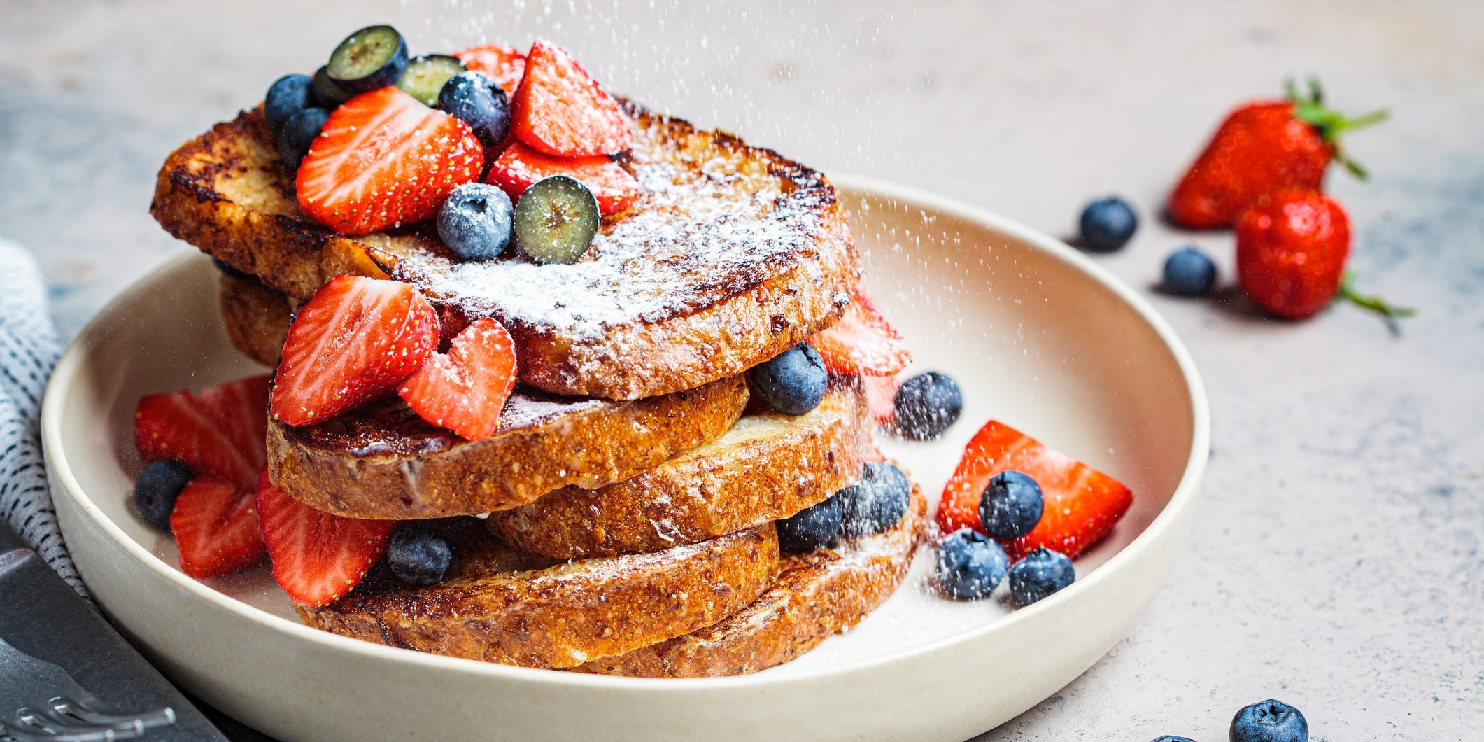 French toast with powdered sugar and berries on top in a white bowl.