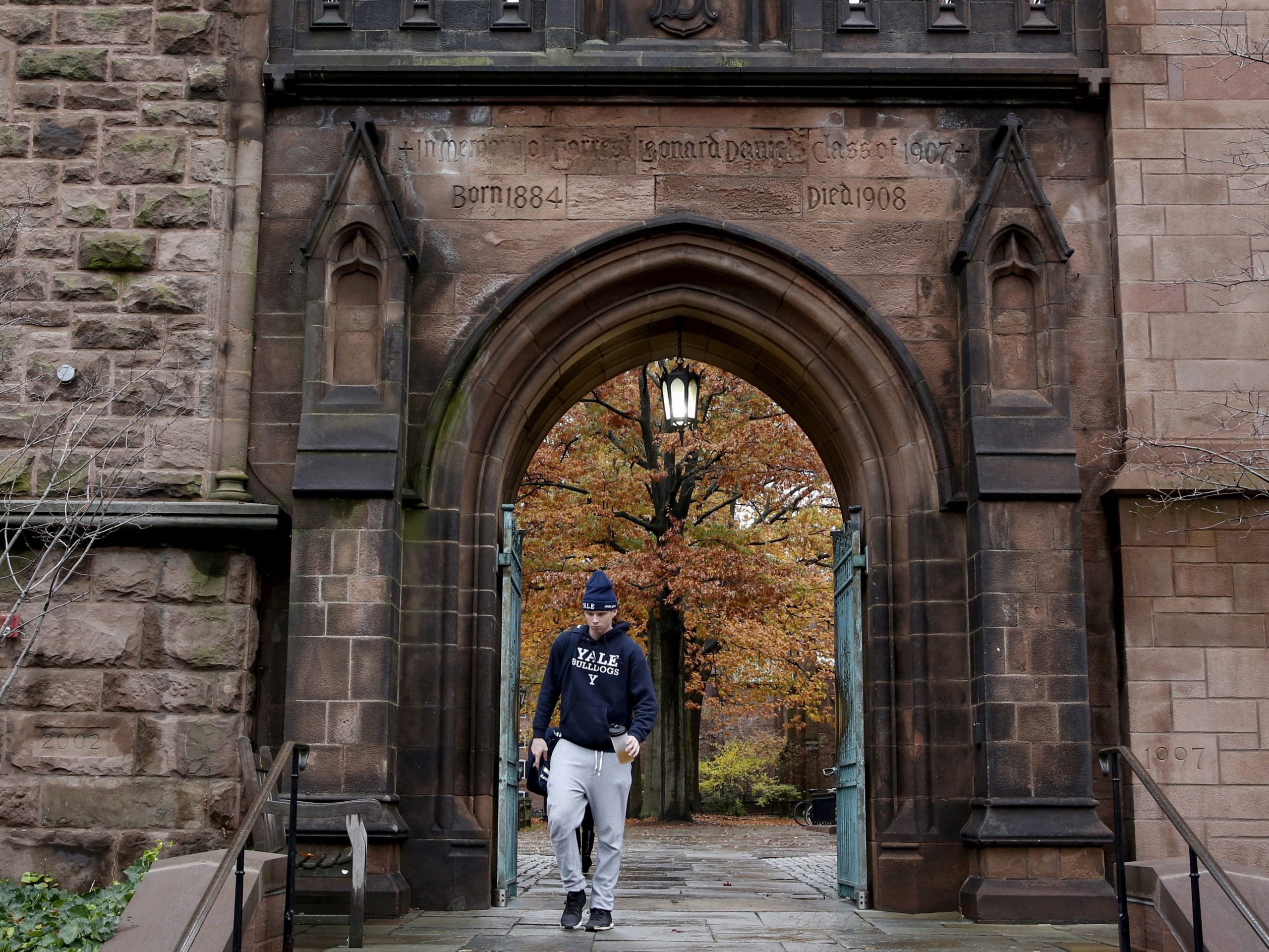A student wearing a sweatshirt and sweatpants walks below a brick archway, with orange and yellow tree leaves in the background.