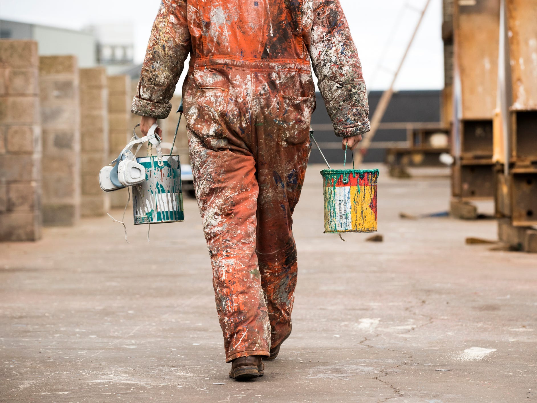 A person in an orange paint-covered jumpsuit carrying two paint cans