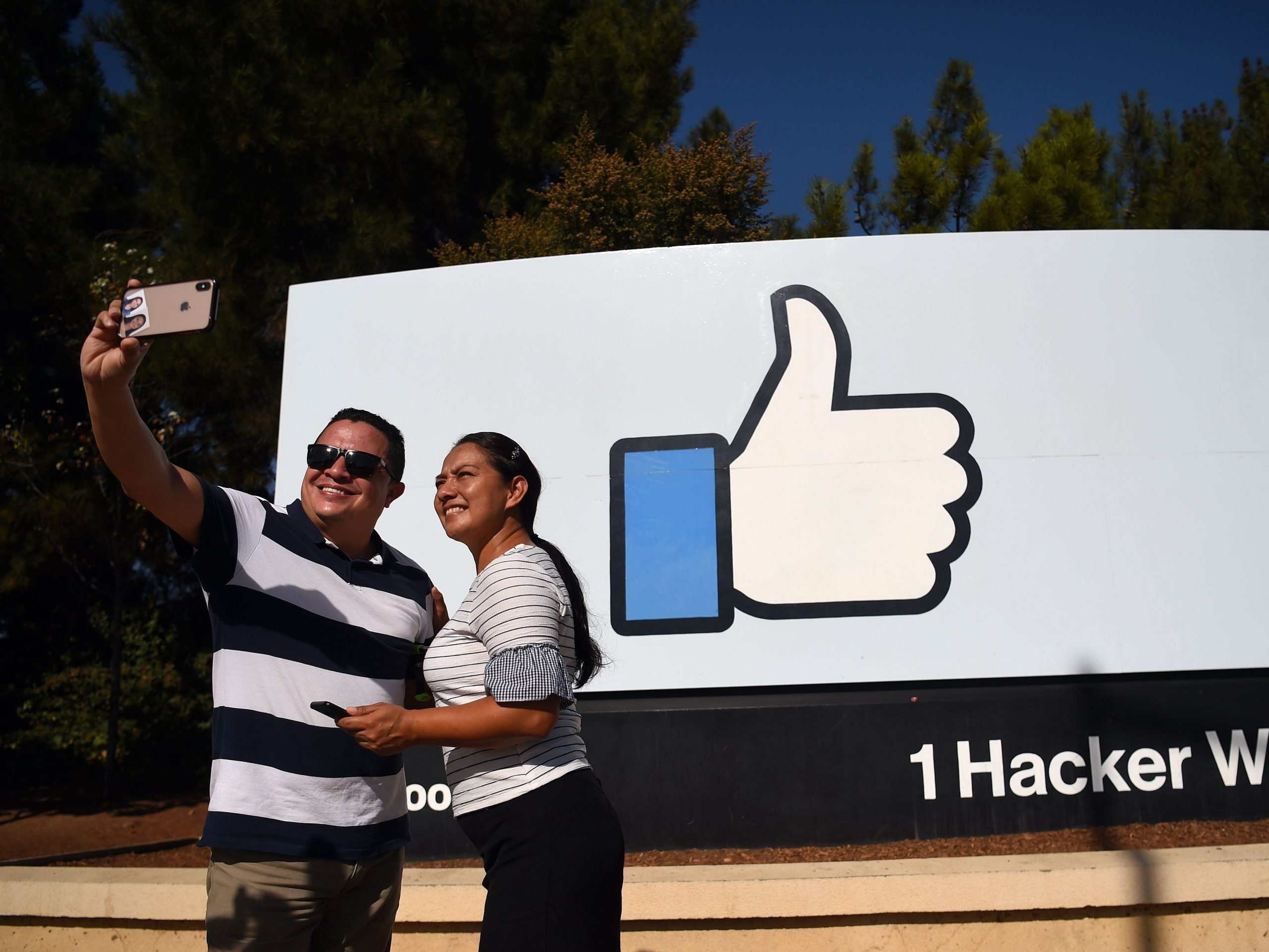 Two people taking a selfie in front of the Facebook "thumbs up" sign at the company's heaequarters