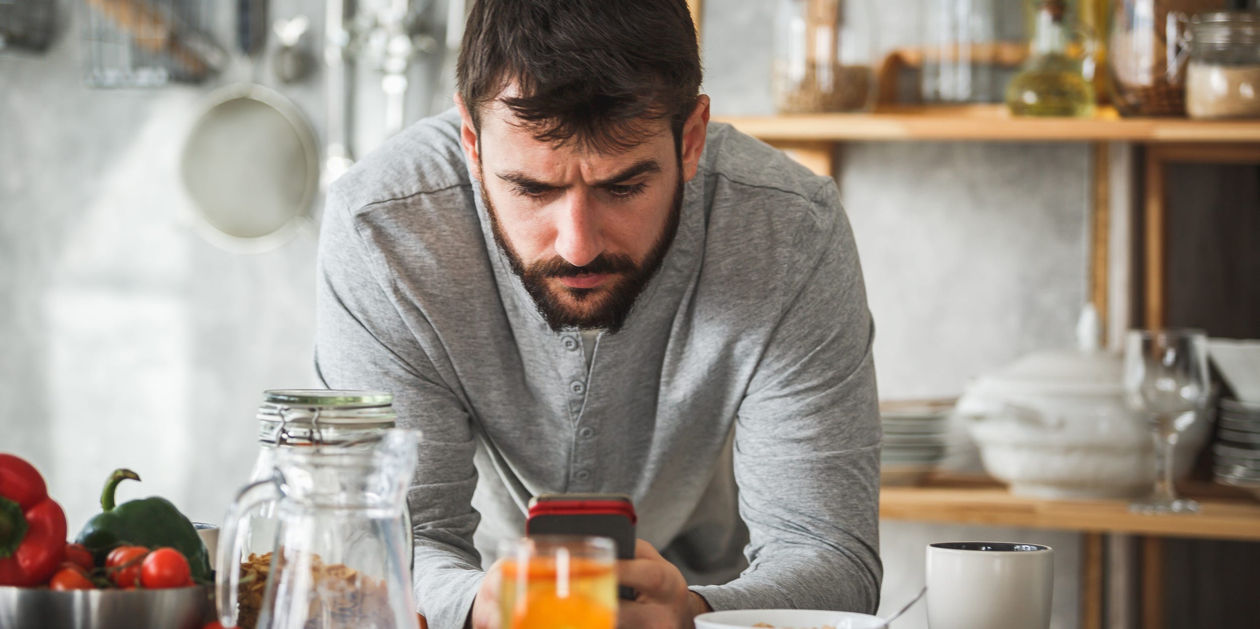 A man frowns at his phone as he stands over a kitchen counter of food.