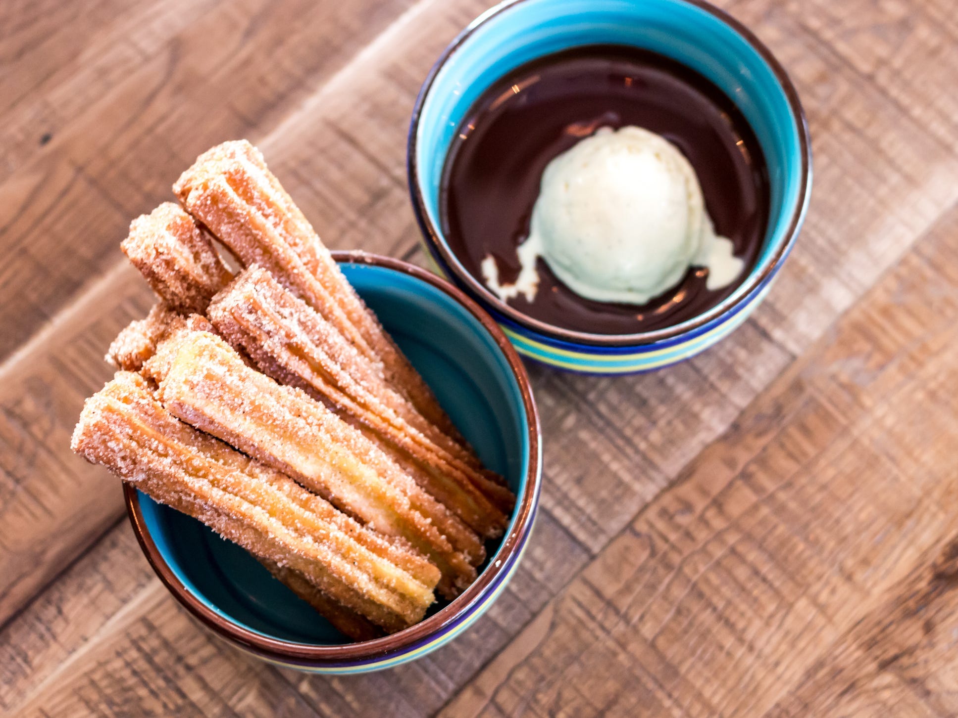 Churros covered in cinnamon sugar stacked vertically in a mug next to a mug filled with chocolate sauce and ice cream