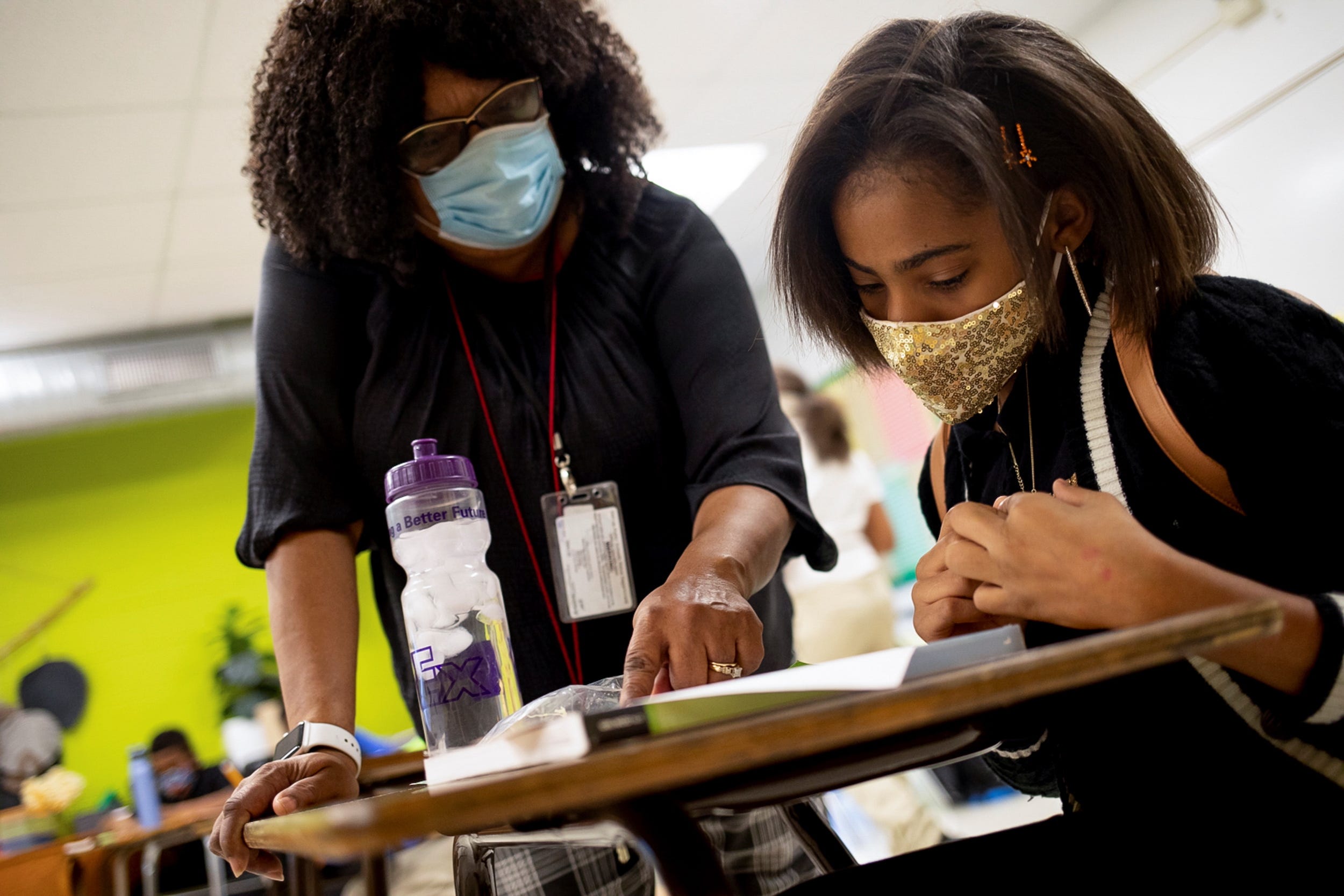 a masked teacher talks to a masked student at her desk in a classroom