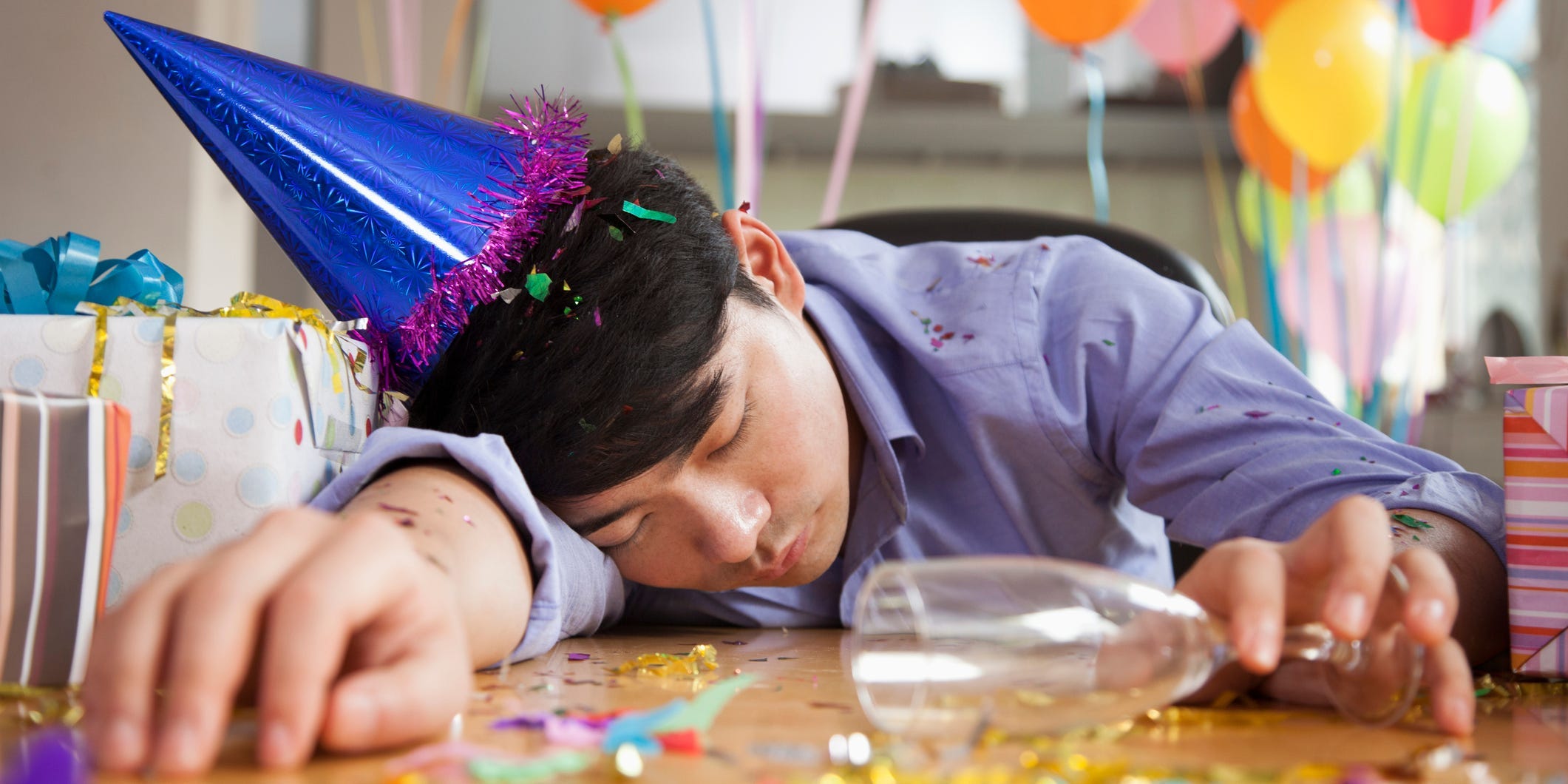 A man in a party hat with balloons behind him lies asleep on a table.