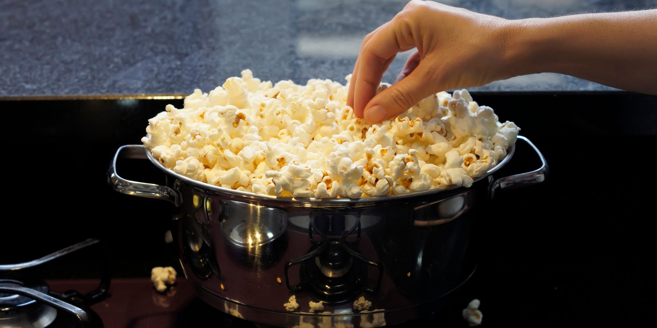 A hand reaching into a pot on the stove overflowing with popcorn