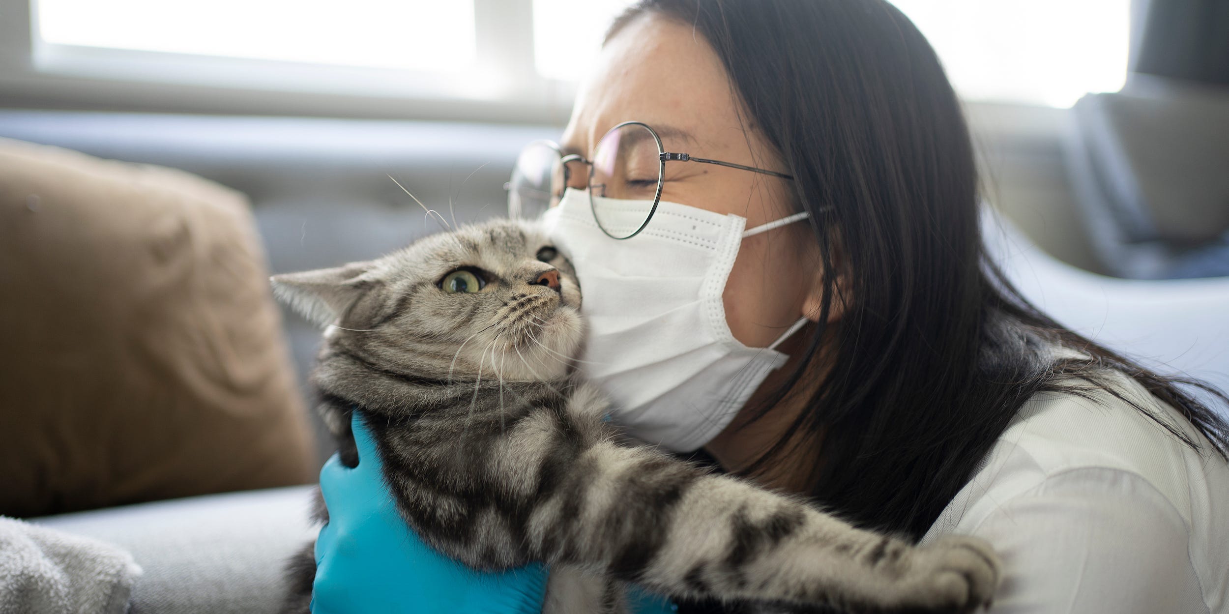 A woman wearing a surgical mask and gloves tries to kiss a cat.