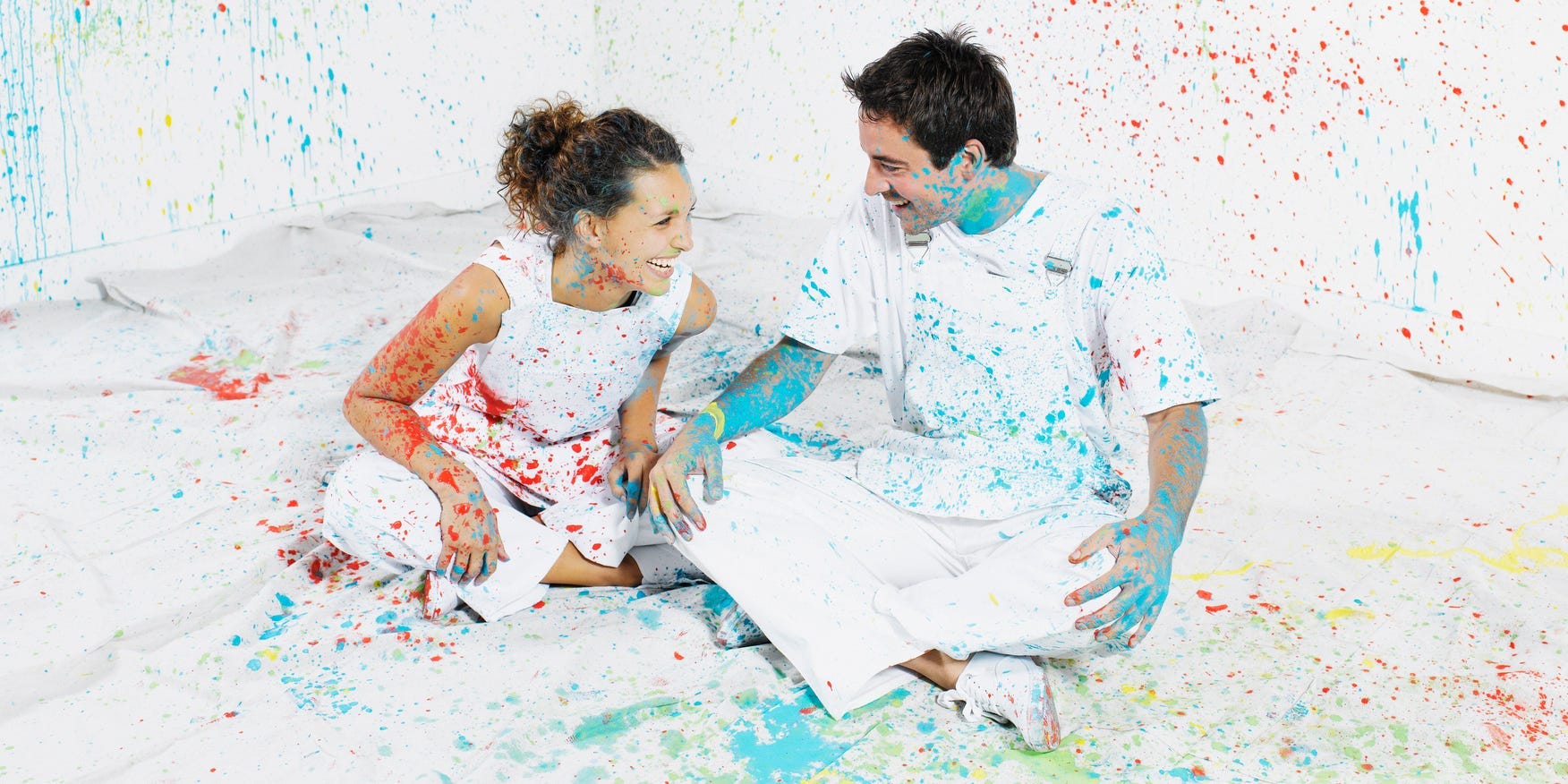 A couple covered in paint sits in a room they splattered with paint.