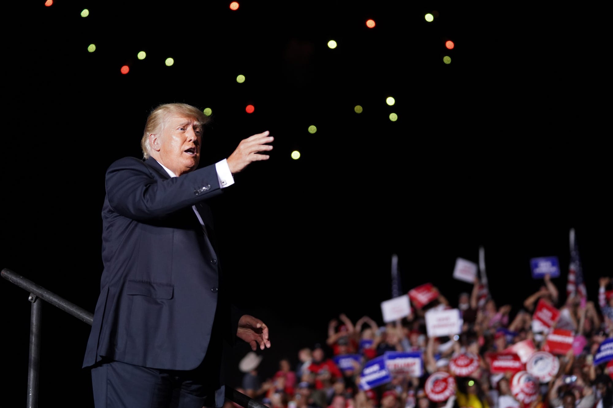 Former President Donald Trump, clad in a dark suit, his right arm extended upward as he stands against a starry sky, waves to attendees at a political rally in Georgia.