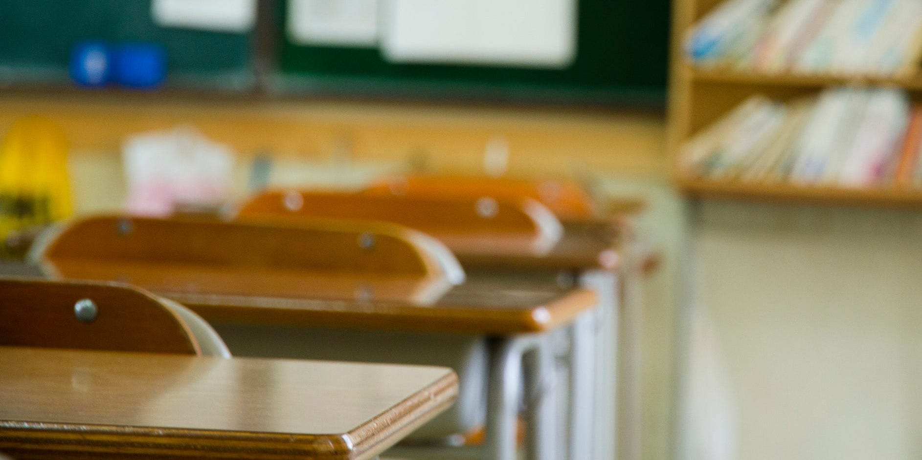 A school classroom, with desks, a blackboard, and bookshelves.