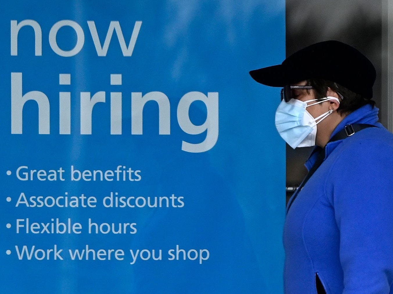 A woman wearing a face mask walks past a "Now Hiring" sign in front of a store.