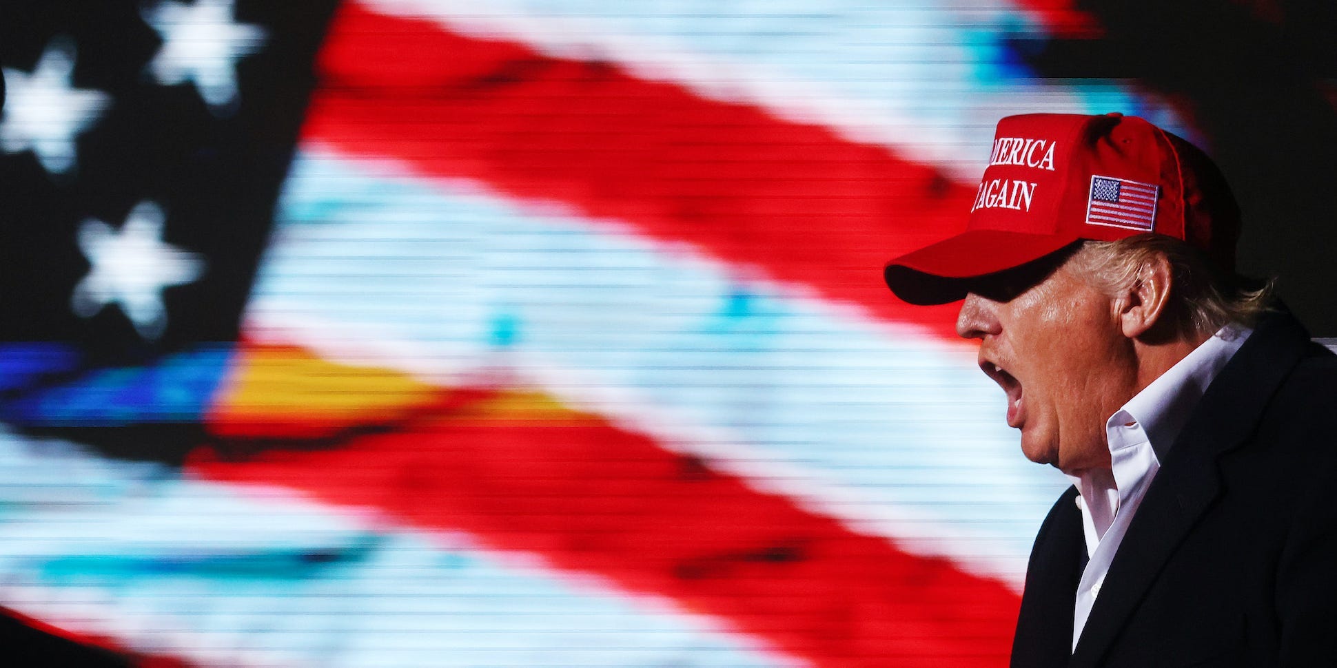 Donald Trump yells in front of an American flag while wearing a MAGA hat.