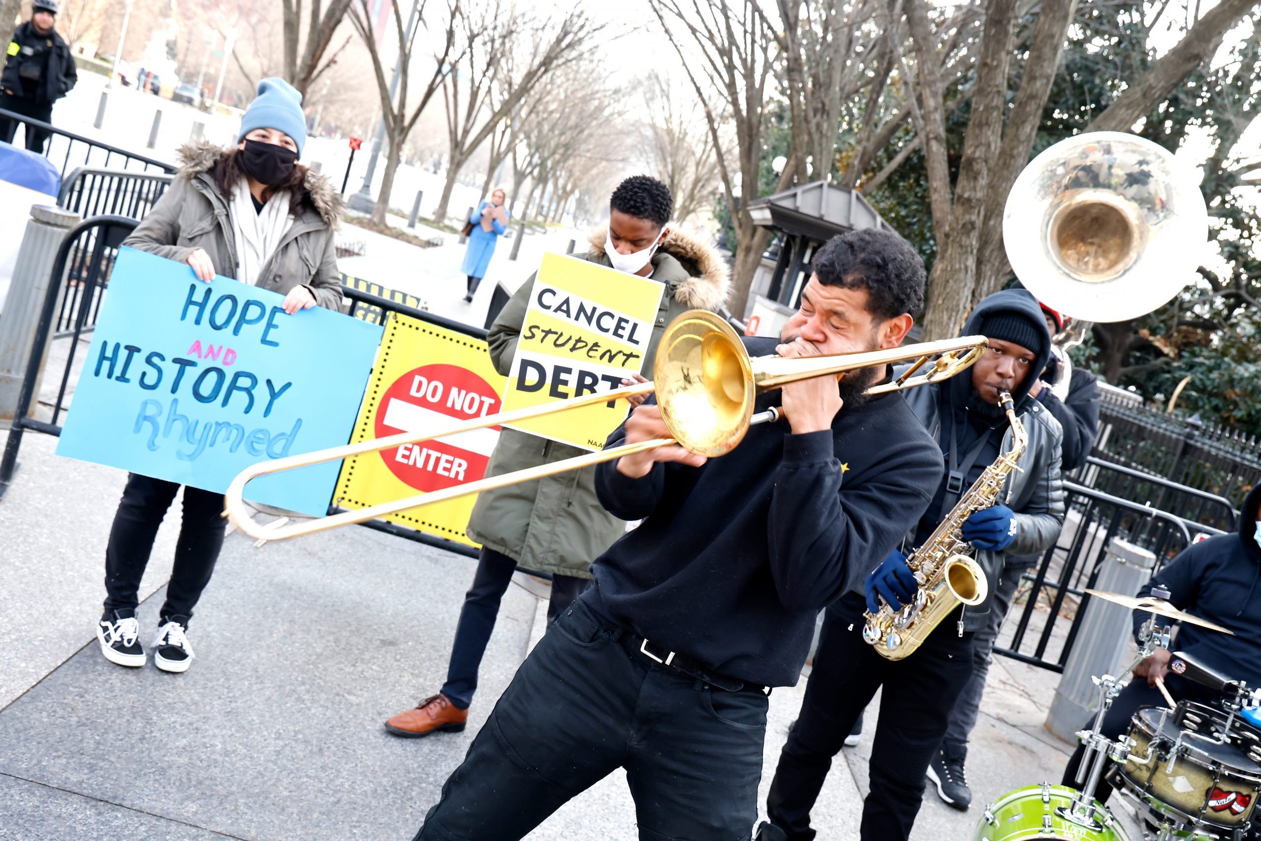 People stand with signs as a man plays the trombone.