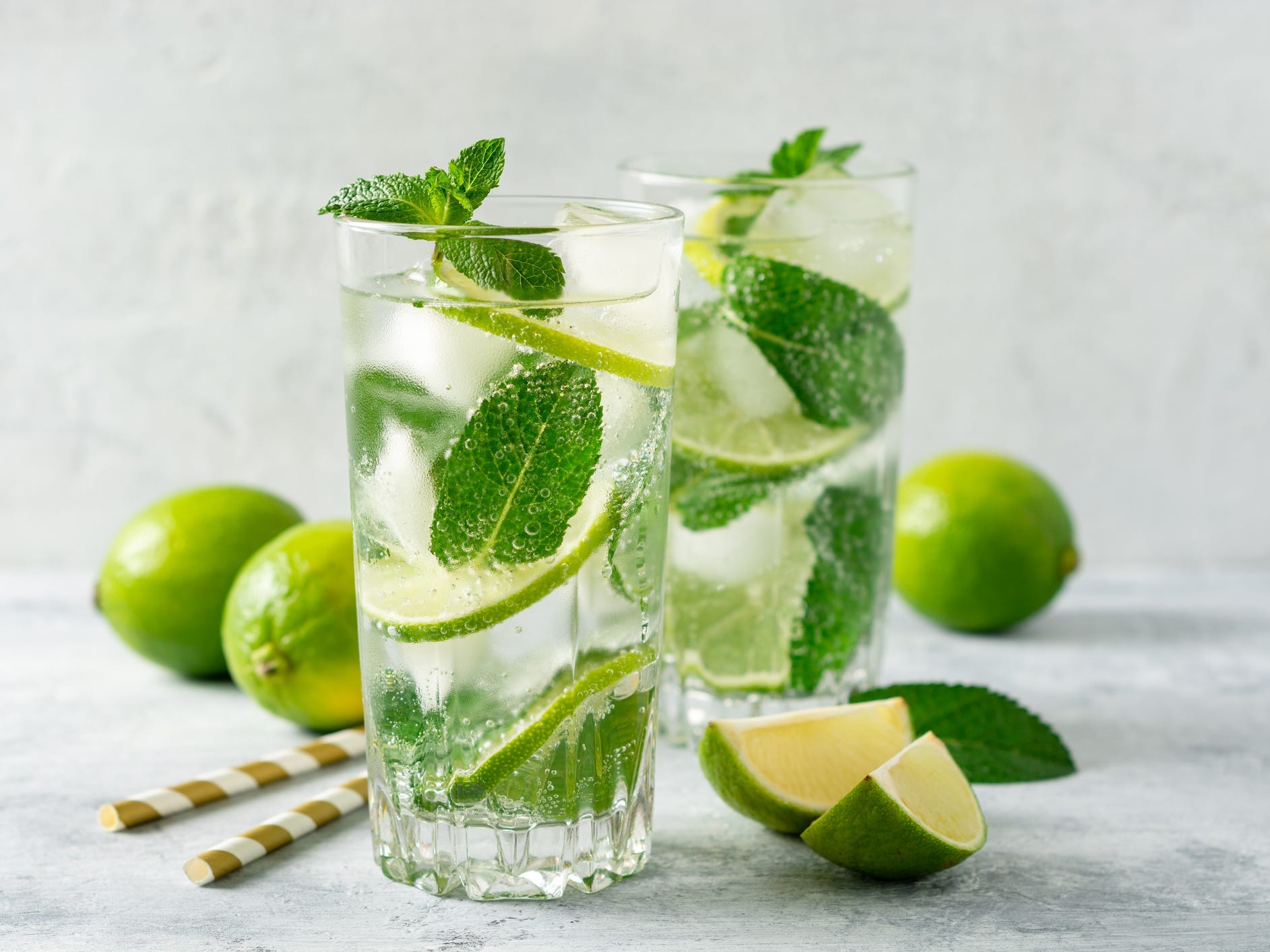 Two moijtos in tall glasses with mint and lime in the glasses