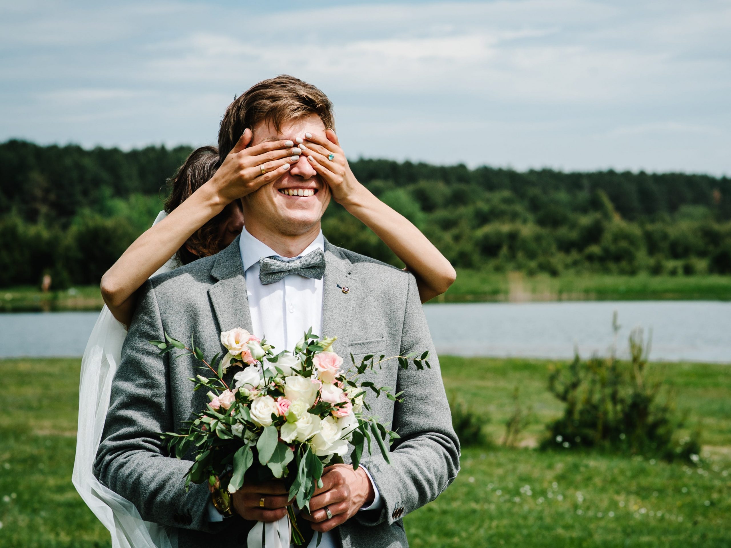 A bride stands behind her groom and covers his eyes before he can see her.