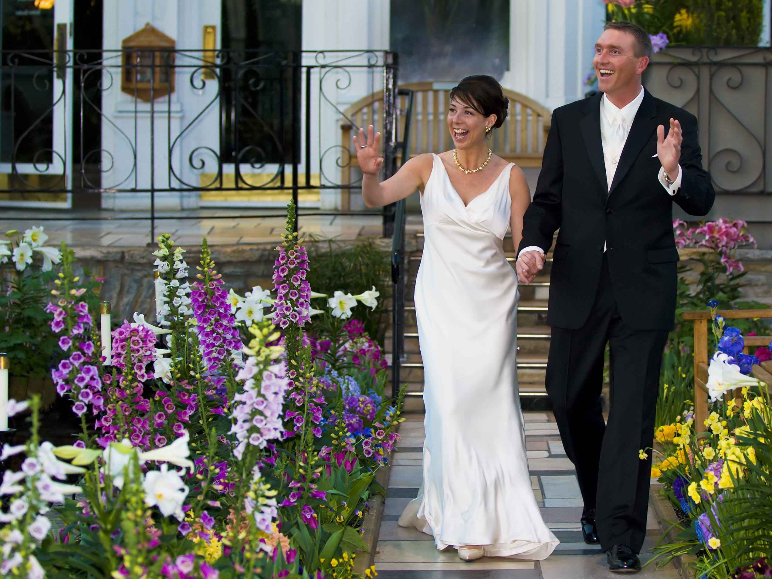 A couple waves as they walk down a less traditional aisle surrounded by flowers.