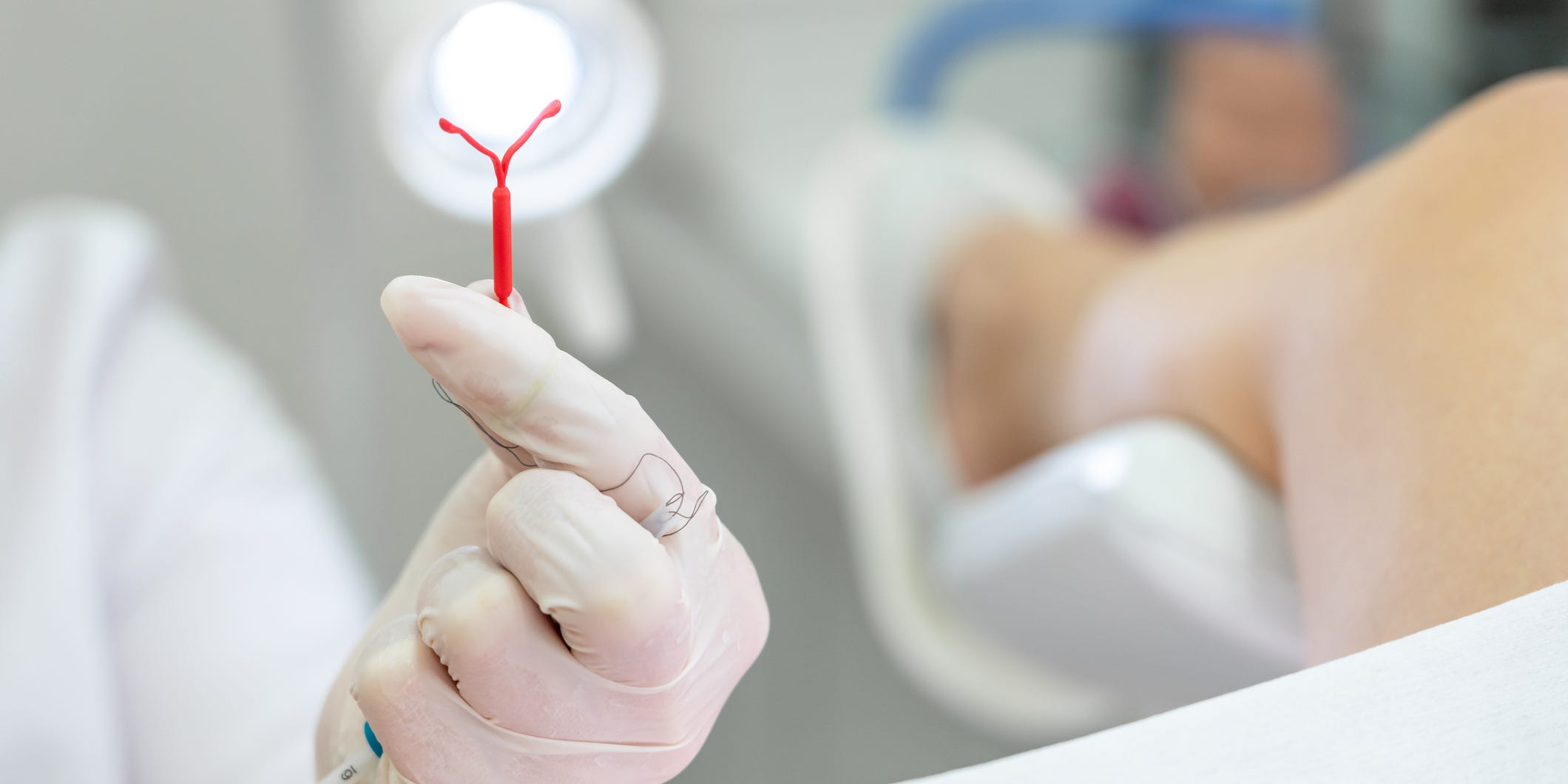 Gynecologist holding an IUD, patient's leg in stirrup in the background.