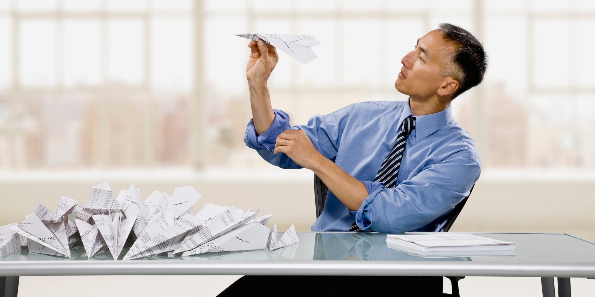 A man sits at his desk procrastinating by making paper airplanes instead of working.