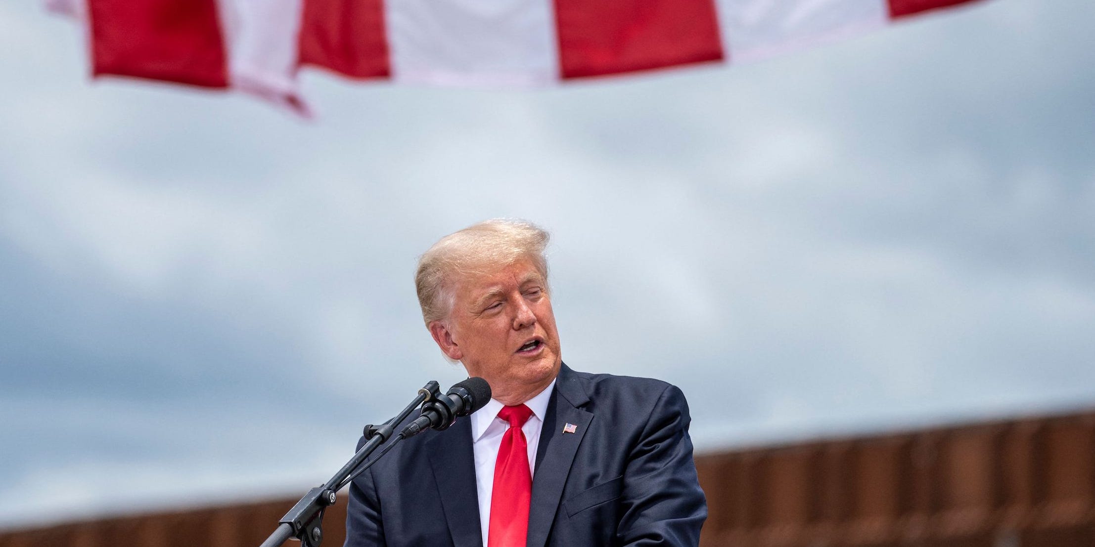 Donald Trump looks to his left under a supersized American flag in front of a partially completed portion of the Mexican border wall.