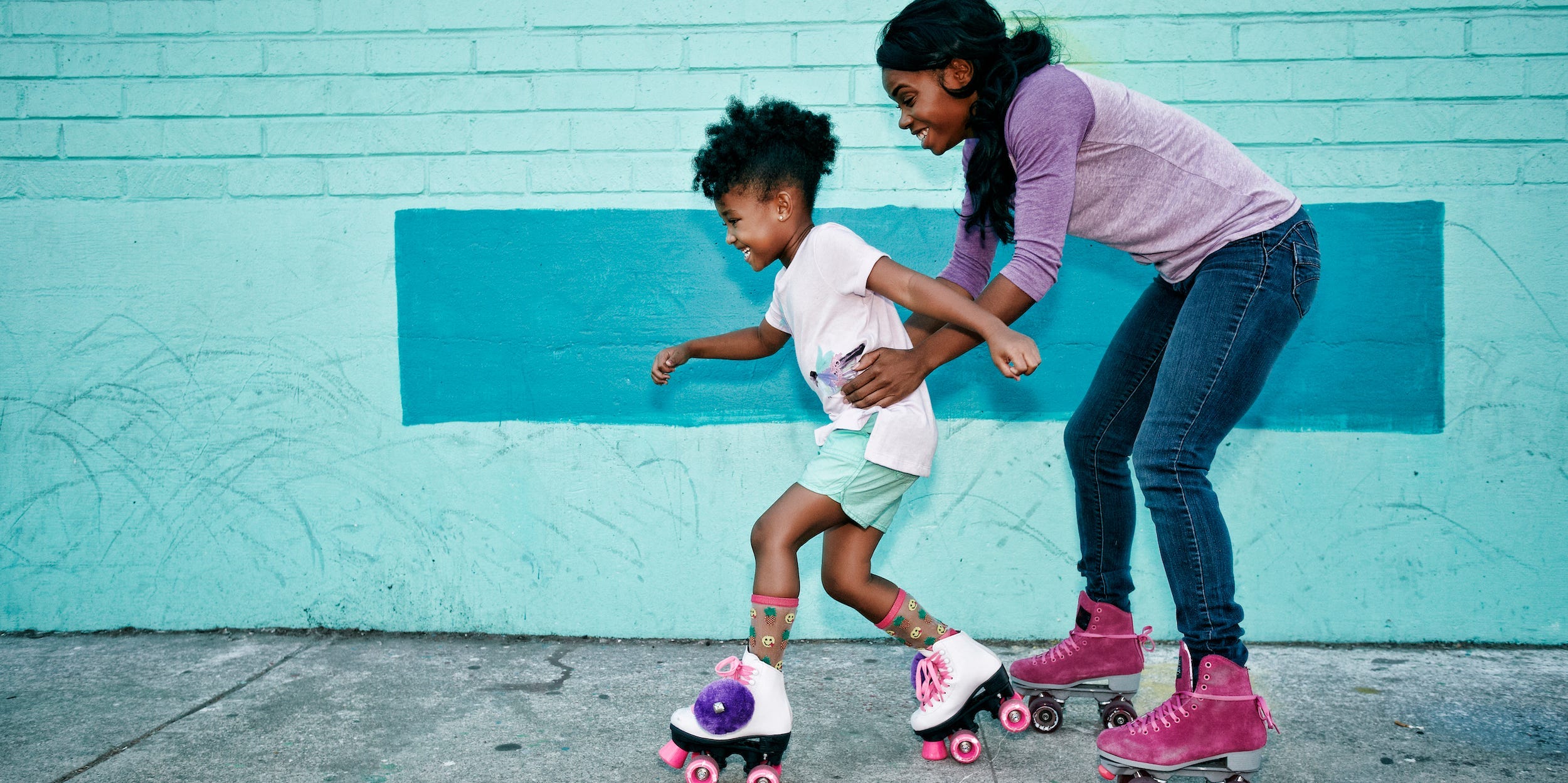A mother supports her daughter's waist as they roller skate.