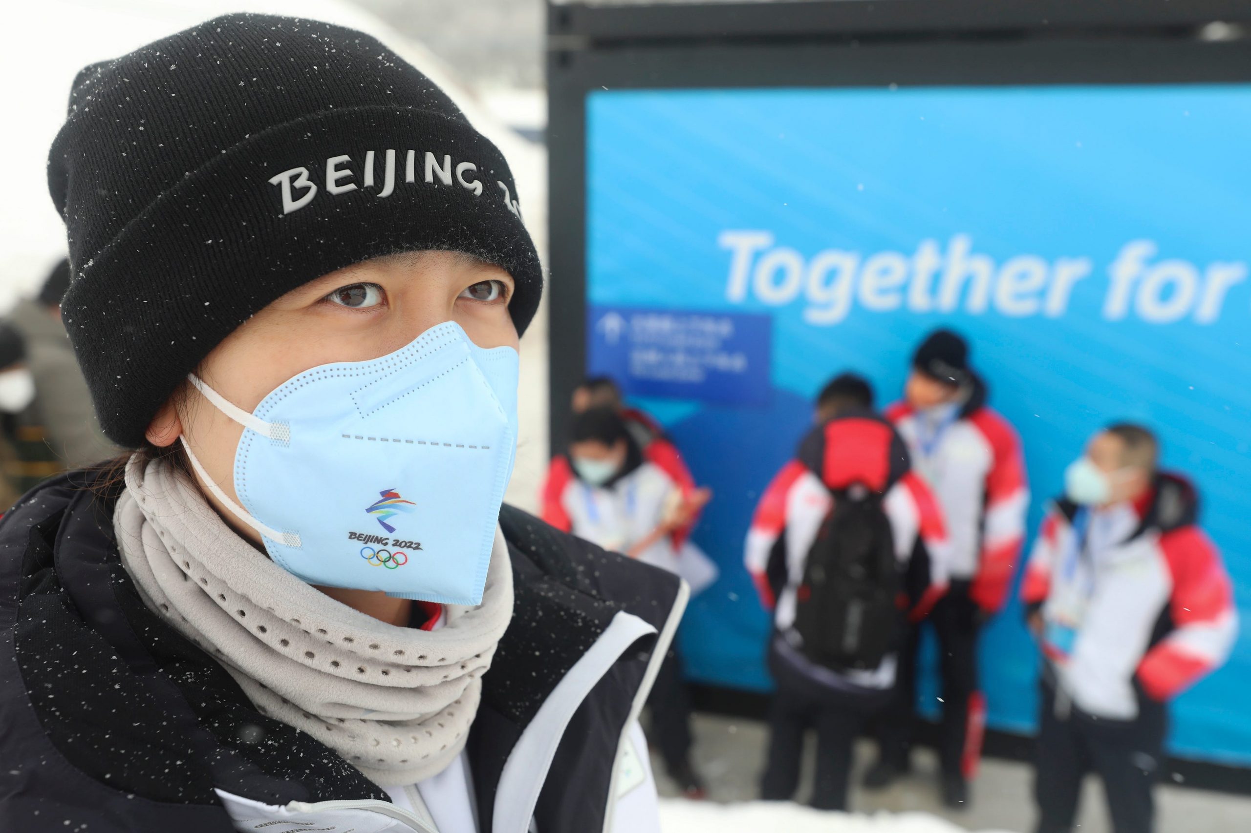 A masked man with a Beijing 2022 Winter Olympics beanie stands in a snow sports facility