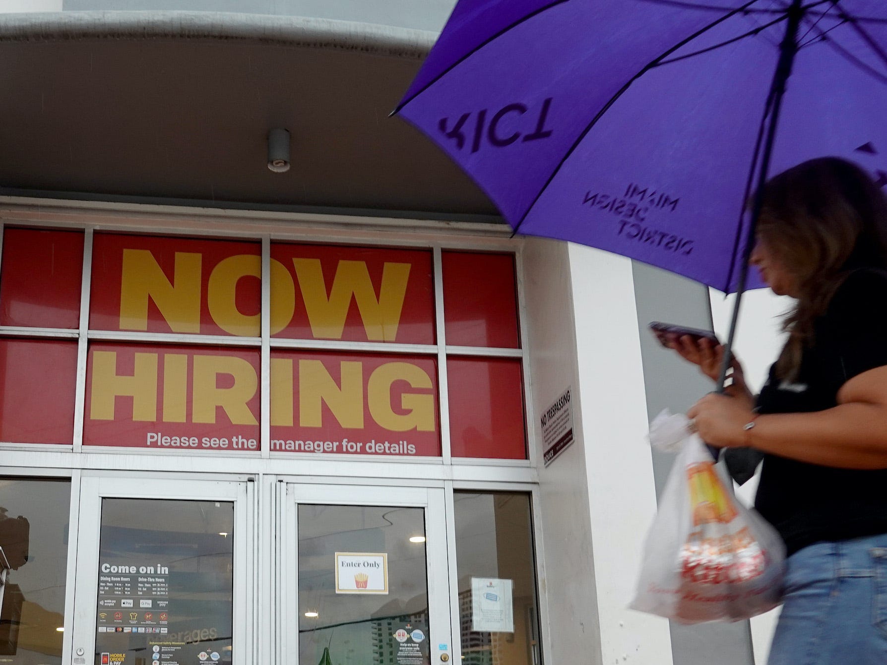 A person with an umbrella walks by a McDonald's restaurant that has a ''Now Hiring" sign above the entrance.