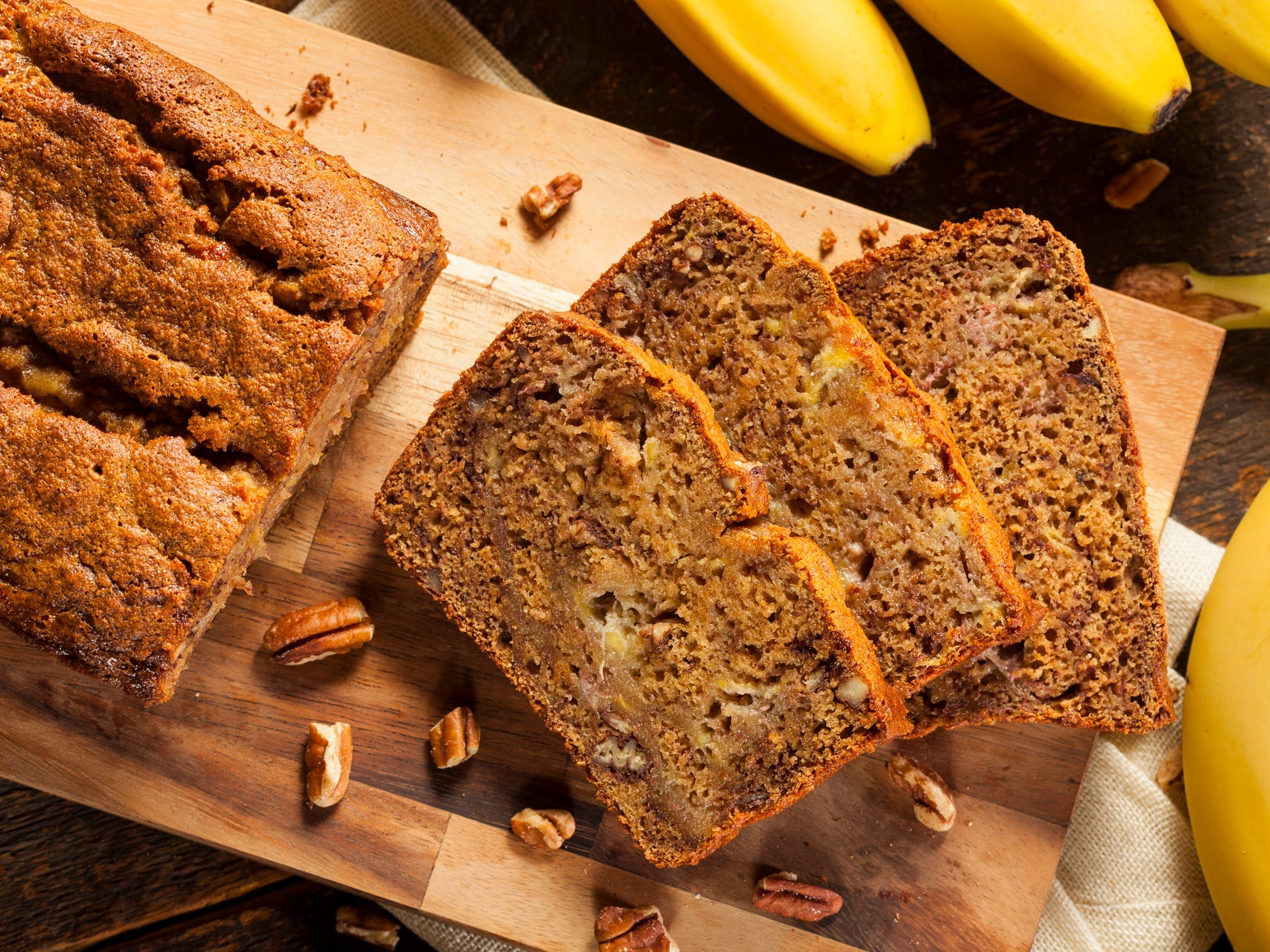 Three slices of banana bread on a cutting board with the rest of the loaf and whole bananas