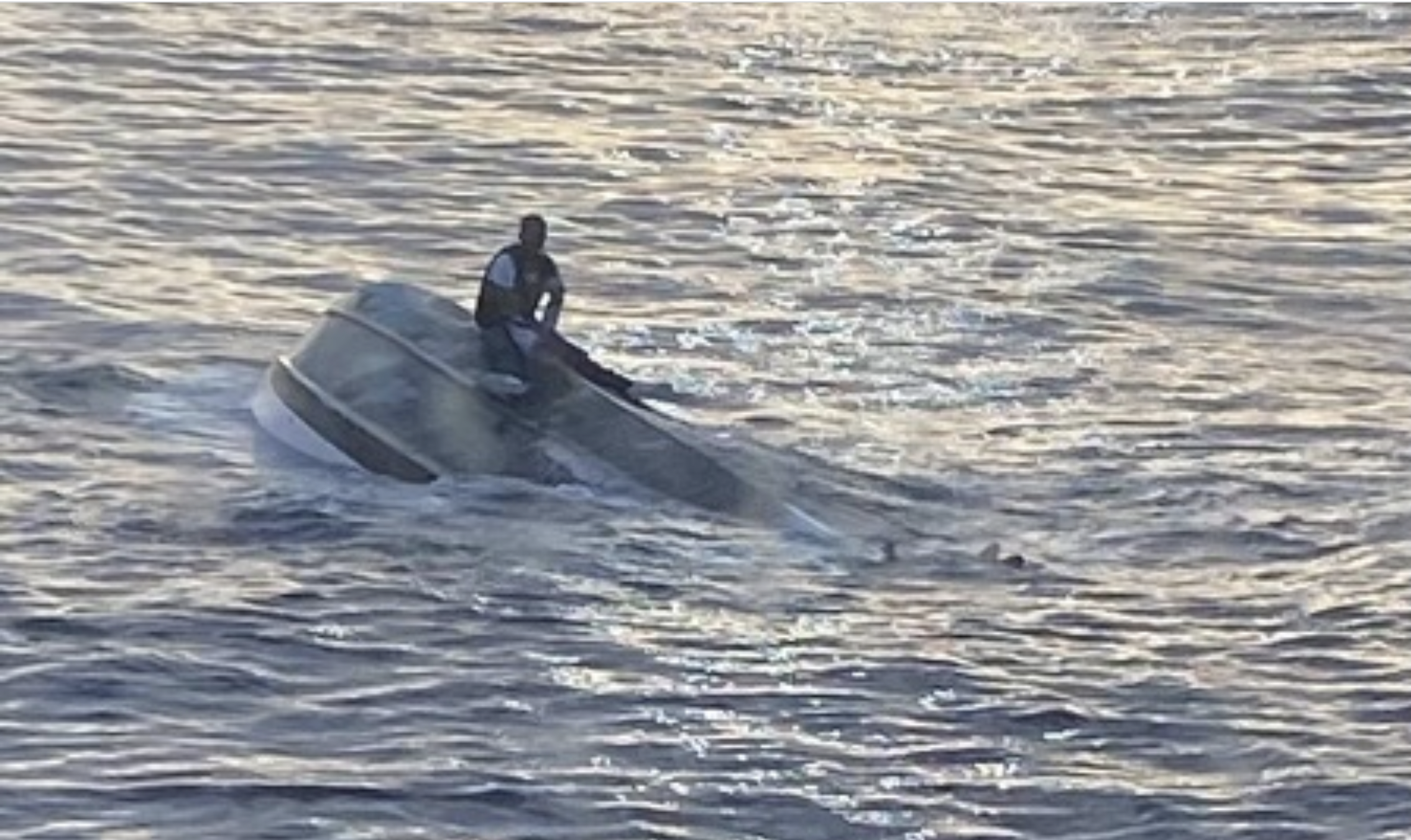 man sitting on top of capsized boat in the middle of water