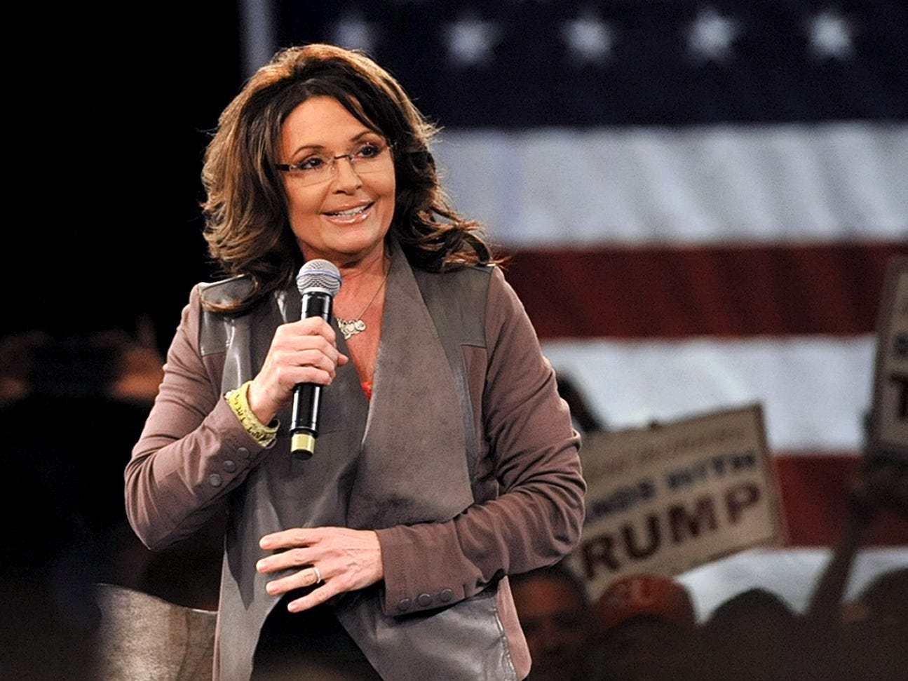 Sarah Pain holds a microphone on stage in front of an American flag and Trump signs