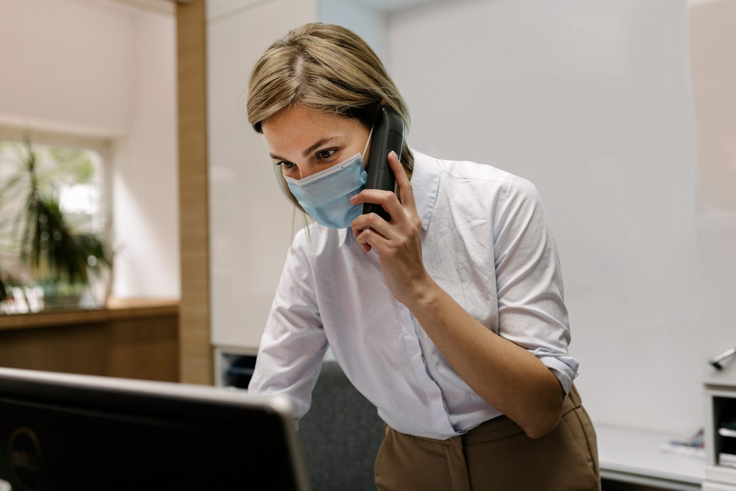 A hotel receptionist following social distancing guidelines and wearing a face mask while working at the front desk and talking on the phone.