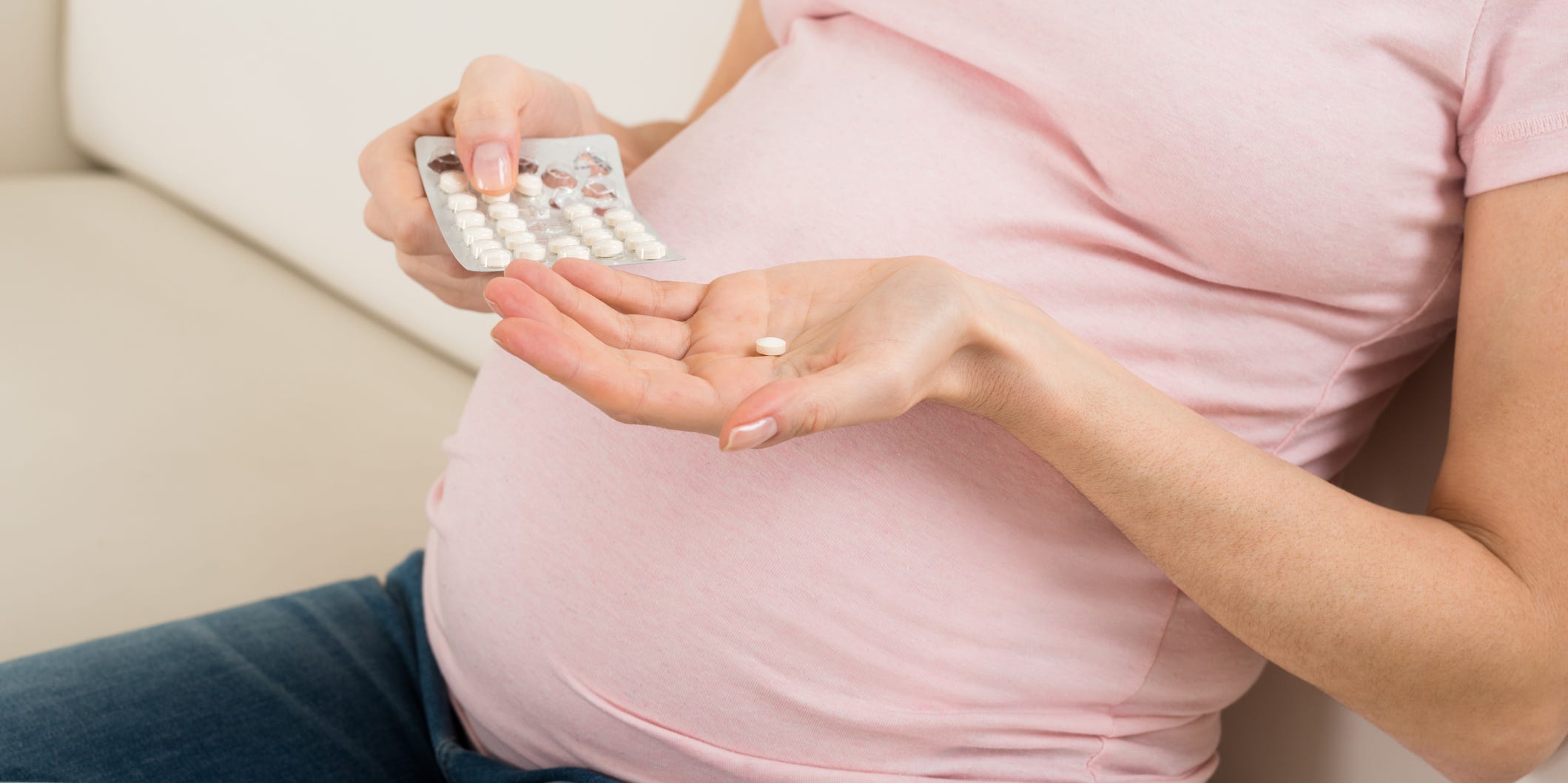 The torso and belly of pregnant woman is pictured. In one hand she holds a pill, in the other she holds the pill package. Over-the-counter medicine like Tylenol.