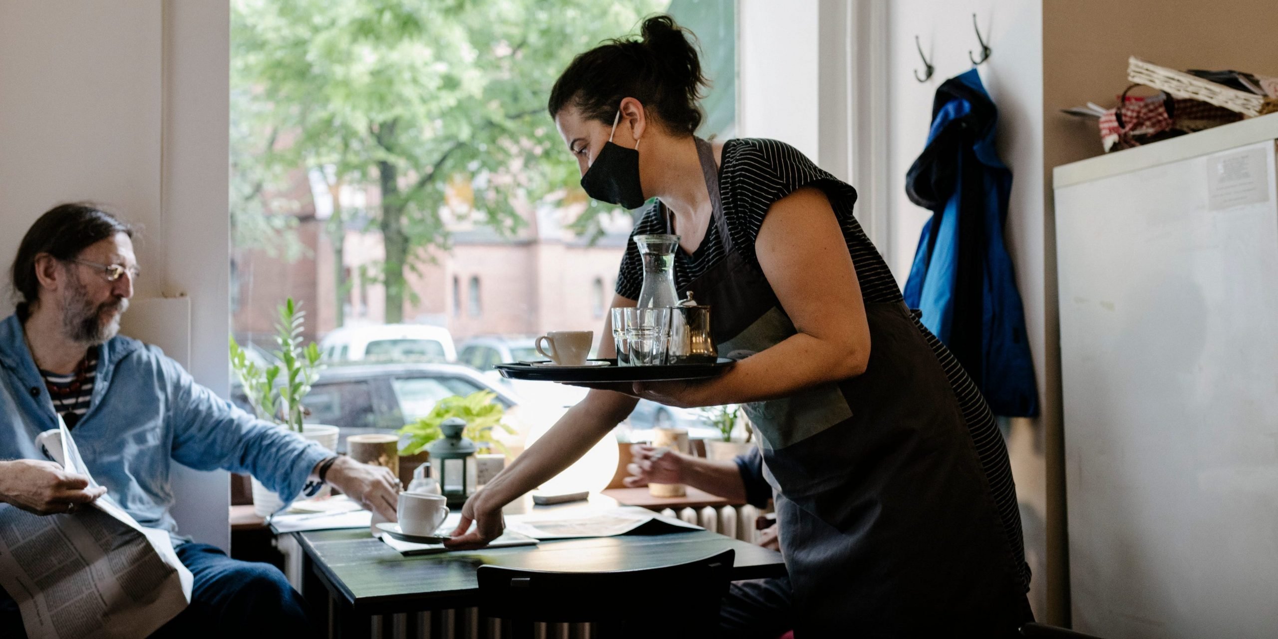 A waitress wearing a face mask and serving a customer some coffee at his table in a restaurant.